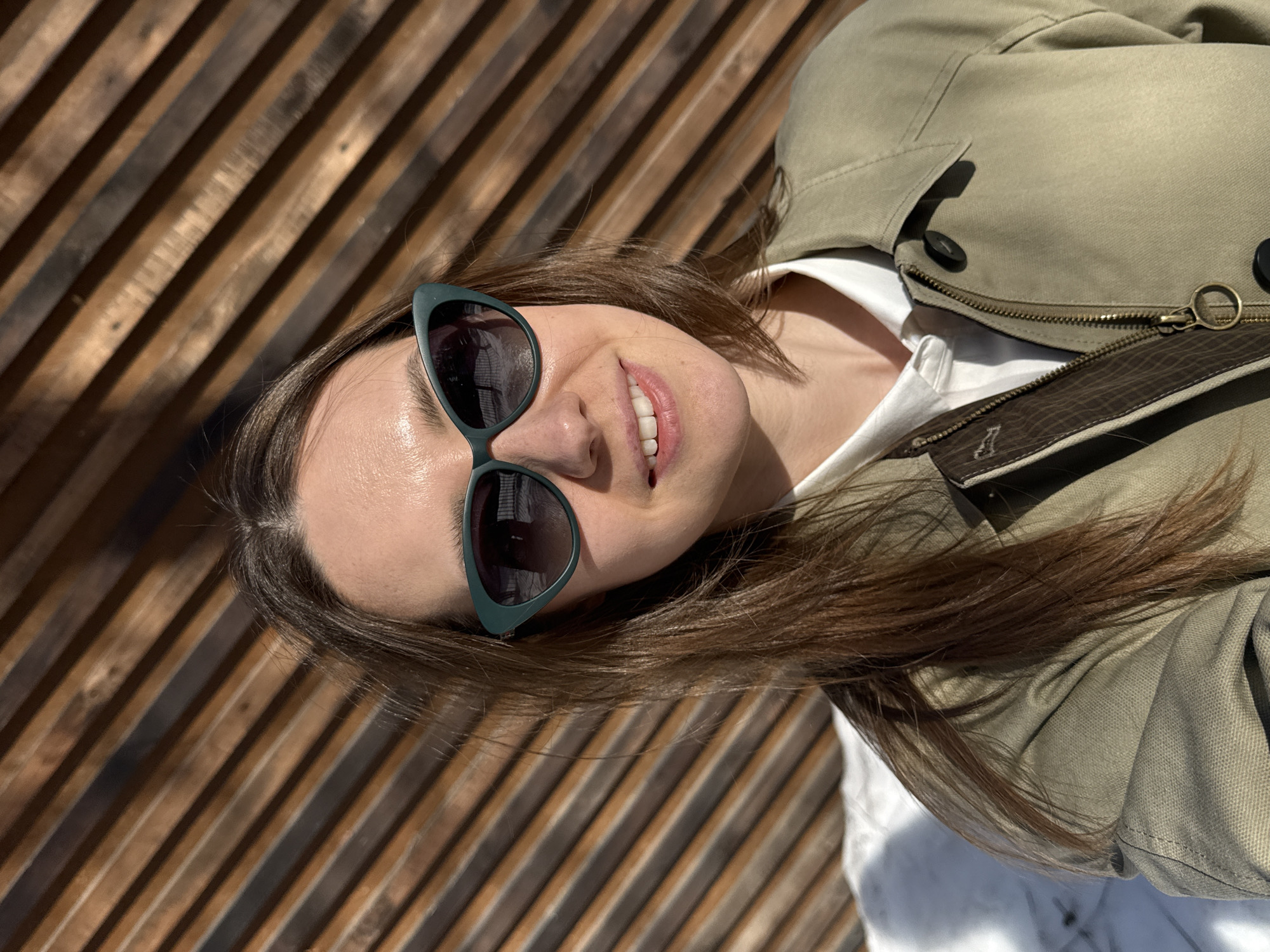 The image is a tilted close-up shot of a woman wearing sunglasses, smiling, and leaning against a wooden structure. The background is made of parallel wooden slats. She is wearing a light-colored jacket over a white collared shirt. The location is Chaikovsky, Russia.