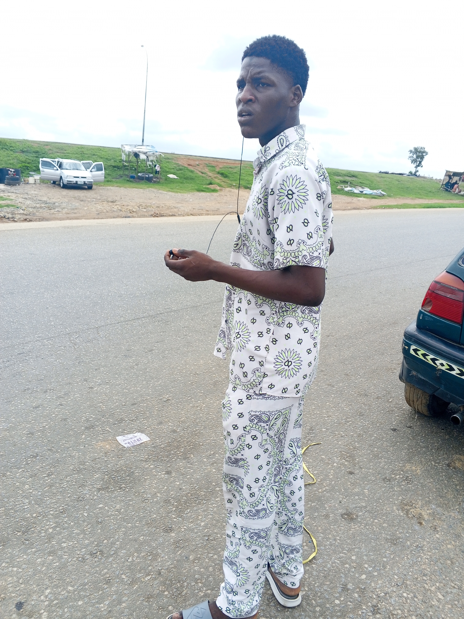 The image shows a young Black male standing on an asphalt road in Pasere, Nigeria. He is wearing a white, patterned shirt and matching pants, and grey sandals. The man is looking to his left, with a slightly concerned expression. He is holding a small black object in his right hand, possibly earbuds. In the background, there is a grassy embankment with a few scattered objects, including a car with its doors open, a makeshift shelter with a light pole, and some tires. To the right of the man, the rear of a dark-colored car is visible. The sky is overcast, suggesting a cloudy day. The scene appears to be outdoors, possibly near a roadside car wash or repair area. A small piece of white paper with visible text is on the road in front of the man. The text on the paper is partially obscured but appears to read "ALLOY".