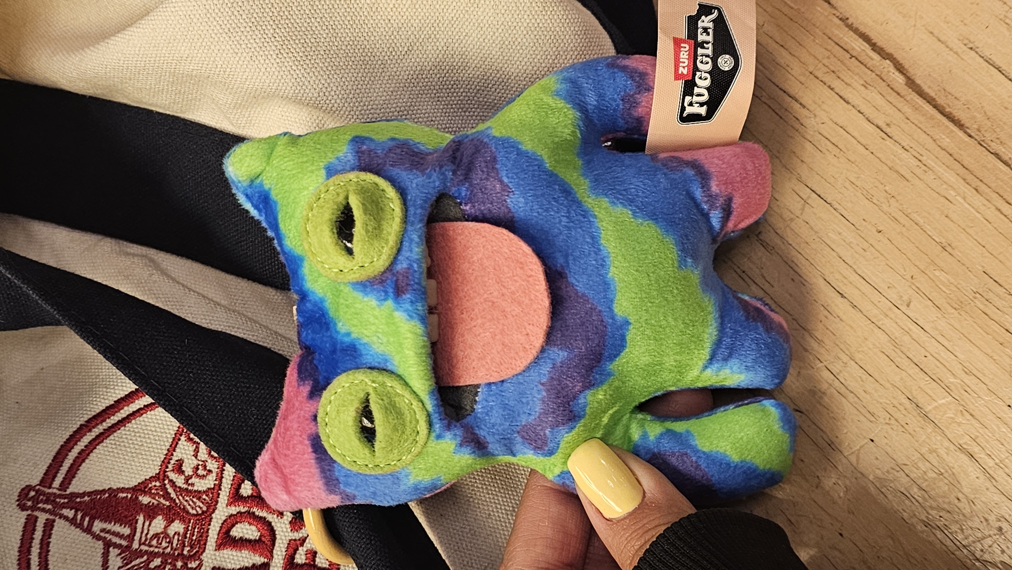 A close-up shot shows a colorful, plush toy, possibly a "Fuggler" brand toy, resting on what appears to be a wooden surface. The toy is a mix of green, blue, purple, and pink hues, featuring large, round, green eyes and a pink tongue. The toy also has a brown "ZURU Fuggler" tag. A hand with yellow painted nails is touching the toy. Behind the toy, there is a partially visible tote bag with a black strap and an embroidered design.
The location is unknown and it is impossible to determine if it is in Puebla, Mexico.