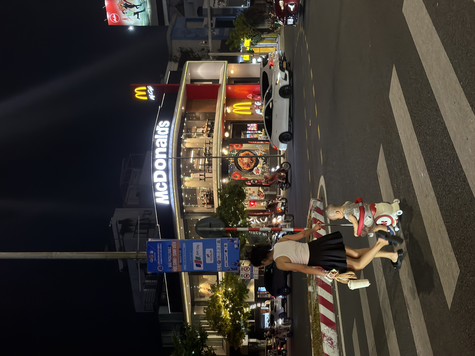 This is a nighttime, medium-angle shot of a McDonald's restaurant in Da Nang, Vietnam. The restaurant is brightly lit with its signature golden arches visible. A woman and a small child are crossing a crosswalk in front of the McDonald's. The woman is wearing a white tank top and a black skirt, and she is holding a bag and a drink. The child is in a red and white stroller. Cars and motorcycles are visible on the street. A tall pole with a blue banner attached to it stands to the left of the McDonald's. The banner has Vietnamese writing and logos. The McDonald's has large windows on the ground floor, showing people inside. There is also a billboard on the right side of the image with a red background and some figures on it. The overall scene depicts a busy urban environment at night.