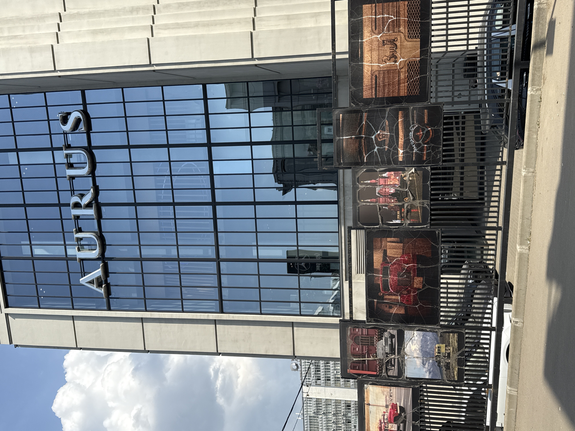 The image shows a building in Moscow, Russia, displaying the Cyrillic script "RUS". The building has a modern architectural style with large glass windows and a concrete exterior. A black metal fence stands in front of the building, displaying images of cars in square frames. A white car is partially visible behind the fence. The sky is partly cloudy with blue patches visible above the building.