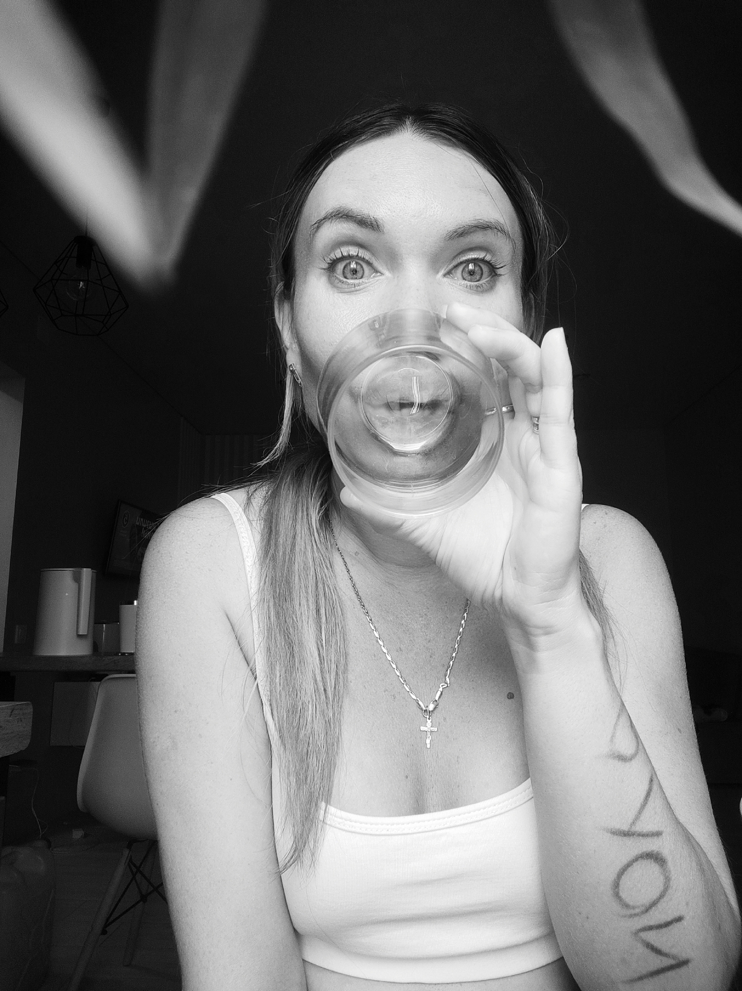 A person is shown in a black and white photograph, holding a clear glass of liquid up to their face. They have wide, expressive eyes and are wearing a white tank top and a silver necklace with a cross pendant. There is writing on their left forearm, and an overhead light fixture is partially visible in the background. The presence of an electric kettle suggests the scene might be inside a home. The location cannot be determined with certainty, the city of Votkinsk cannot be determined with this image.