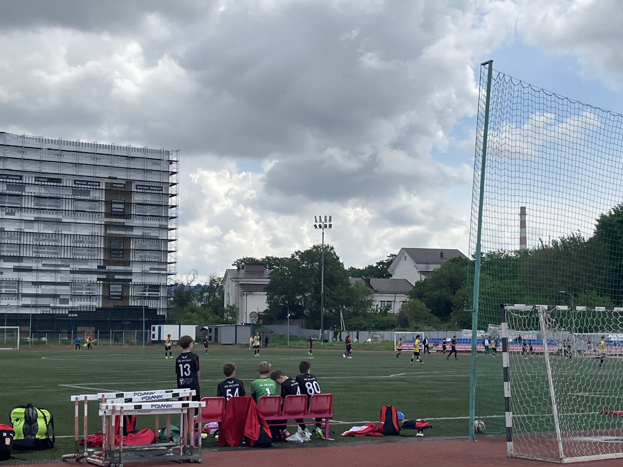 A soccer game is in progress on a green artificial turf field, likely in Krasnodar, Russia, based on the architectural style of nearby buildings. Several young players in yellow and black uniforms are actively playing. Near the sideline, four boys are sitting on a red bench, facing the field; they wear soccer jerseys with the numbers 13, 54, 1, and 80. Gear bags are nearby. The background includes a multi-story building under construction with scaffolding and several other buildings nestled among trees. A tall lighting structure is visible in the middle ground. A soccer goal with a net is in the foreground. The sky is overcast with heavy clouds. The foreground also features hurdles and sports equipment beside the bench.