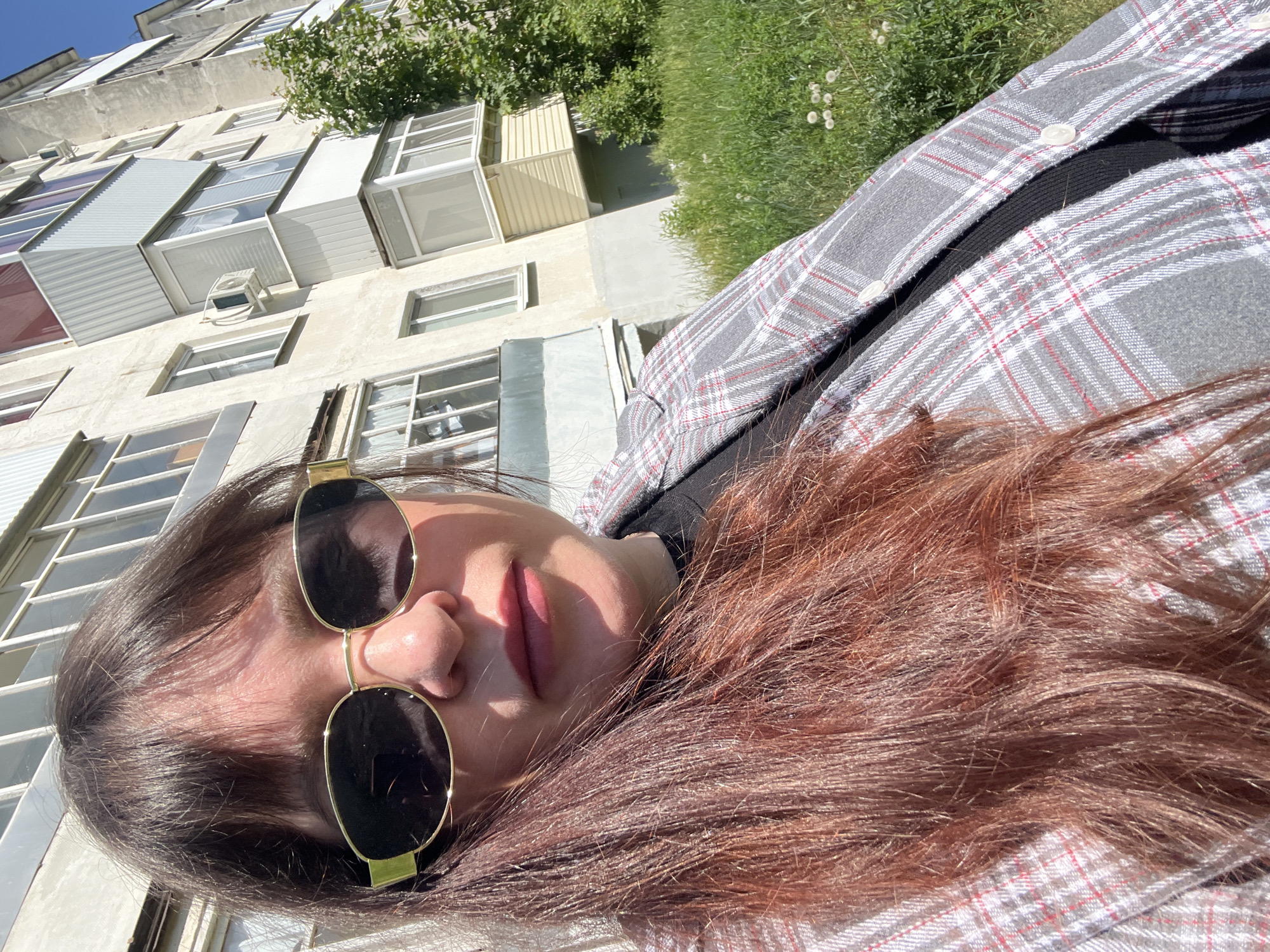 A selfie of a woman with long brown hair and sunglasses, wearing a gray and white plaid shirt over a black top. The background shows a multi-story building with protruding, irregularly shaped windows and air conditioning units. Greenery is visible above the building. The location is Noviy, Russia.