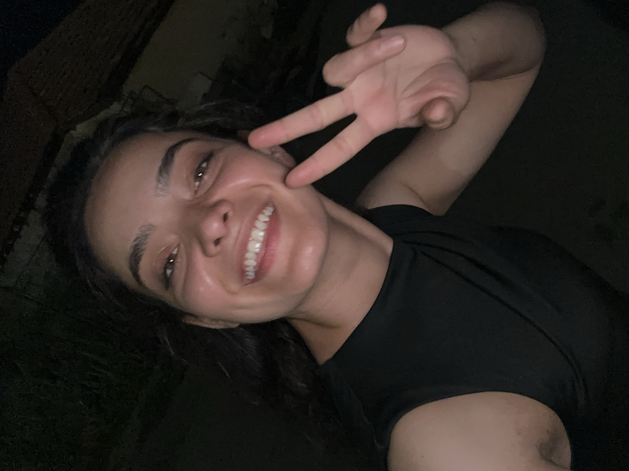 A young woman of Eastern European descent, with dark hair pulled back and a bright smile, is captured in a close-up selfie taken at night. She is wearing a black sleeveless top. Her right hand is raised, forming a peace sign, with her index and middle fingers extended. The background is dark and out of focus, suggesting an outdoor setting. The image appears to have been taken with a smartphone in a low-light environment, resulting in a slightly grainy texture. The location is stated to be Malevo, Bulgaria.