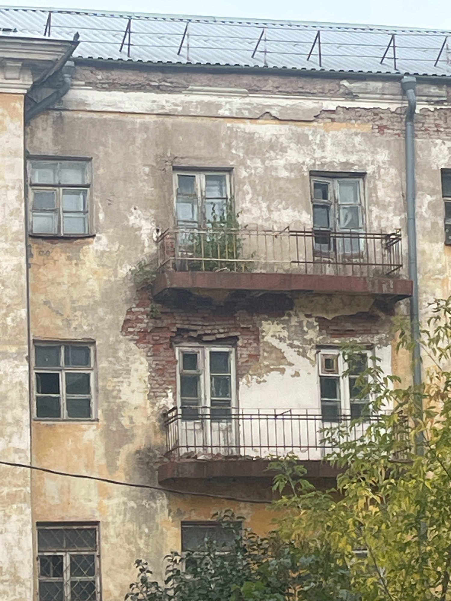 The image depicts the exterior of an aged, multi-story building in Sof'ino, Russia. The building exhibits signs of deterioration, with peeling paint, exposed brickwork, and weathered facades. There are multiple windows visible across the different levels, some of which appear to be old-fashioned with multiple panes. Two balconies are present, each featuring a rusted metal railing. Green foliage, likely trees and bushes, partially obscures the lower portions of the building and frames the scene on the right side. The sky above is overcast.