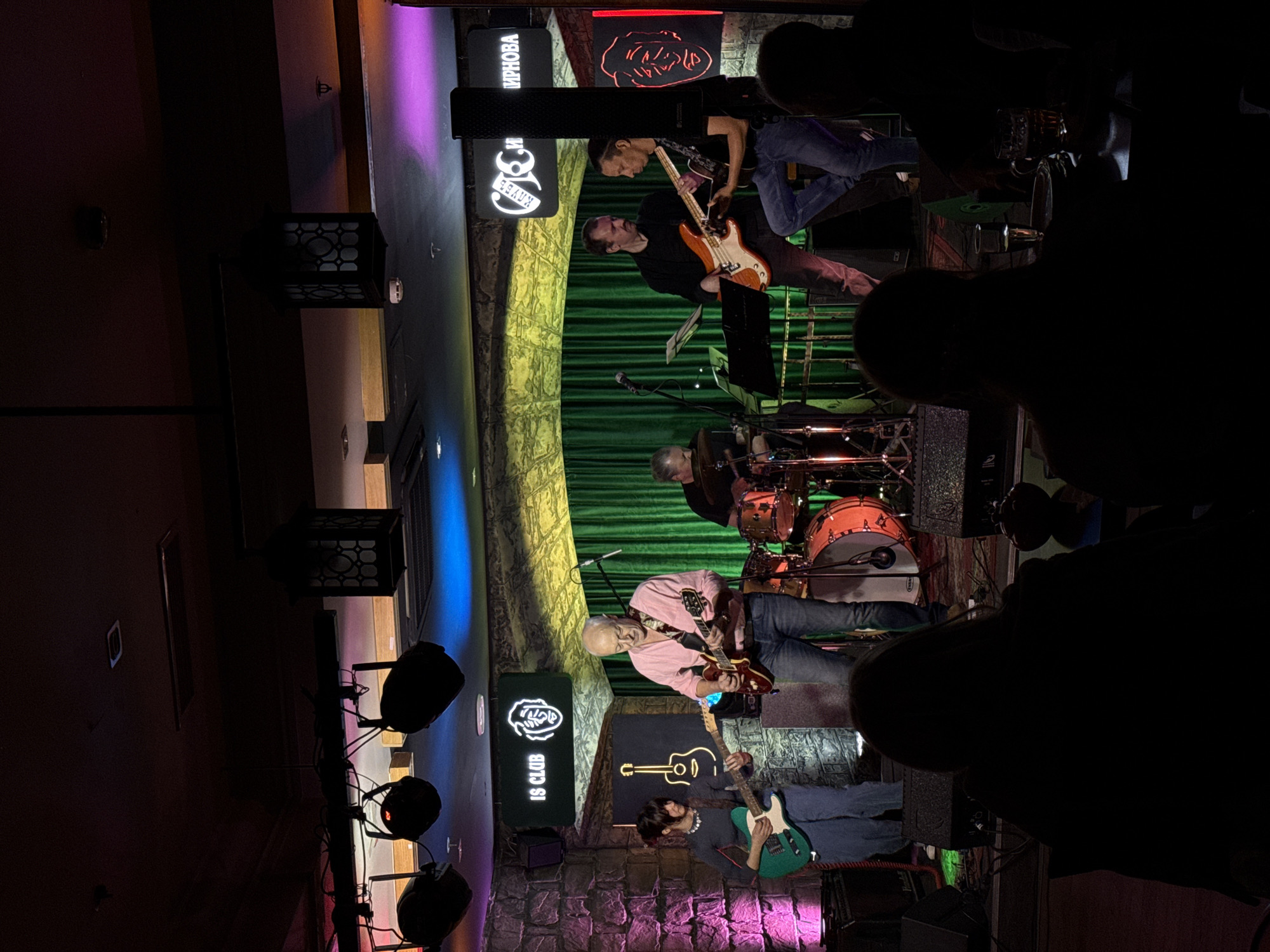This image captures a live musical performance by a band on a stage inside a club, likely in Moscow, Russia, during the evening or night. The scene is dimly lit with vibrant stage lights casting purple, green, and orange hues across the performers and the backdrop.
There are at least four visible musicians on stage. In the center, an older man with white hair and a beard, wearing a light pink shirt and blue jeans, is seated on a stool playing a red electric guitar. To his right, a man in a black shirt is standing and playing an orange electric guitar or bass. Behind them, partially obscured, a drummer is visible with a drum set featuring orange-toned drums. To the far left, another musician, wearing a dark shirt and blue jeans, is standing and playing a green electric guitar. Microphones and music stands are present on the stage.
The stage background consists of rich green curtains and a textured wall that appears to be made of stone. Above the stage, several signs are visible. One sign clearly shows "КЛУБ" (CLUB) in Cyrillic, with "IS CLUB" in English directly below it. Another sign features a stylized illustration of a head or face.
The foreground of the image is dominated by the dark silhouettes of audience members, indicating they are standing and watching the concert. The overall ambiance is characteristic of a music venue, with the colorful stage lighting creating a focal point on the band and instruments.