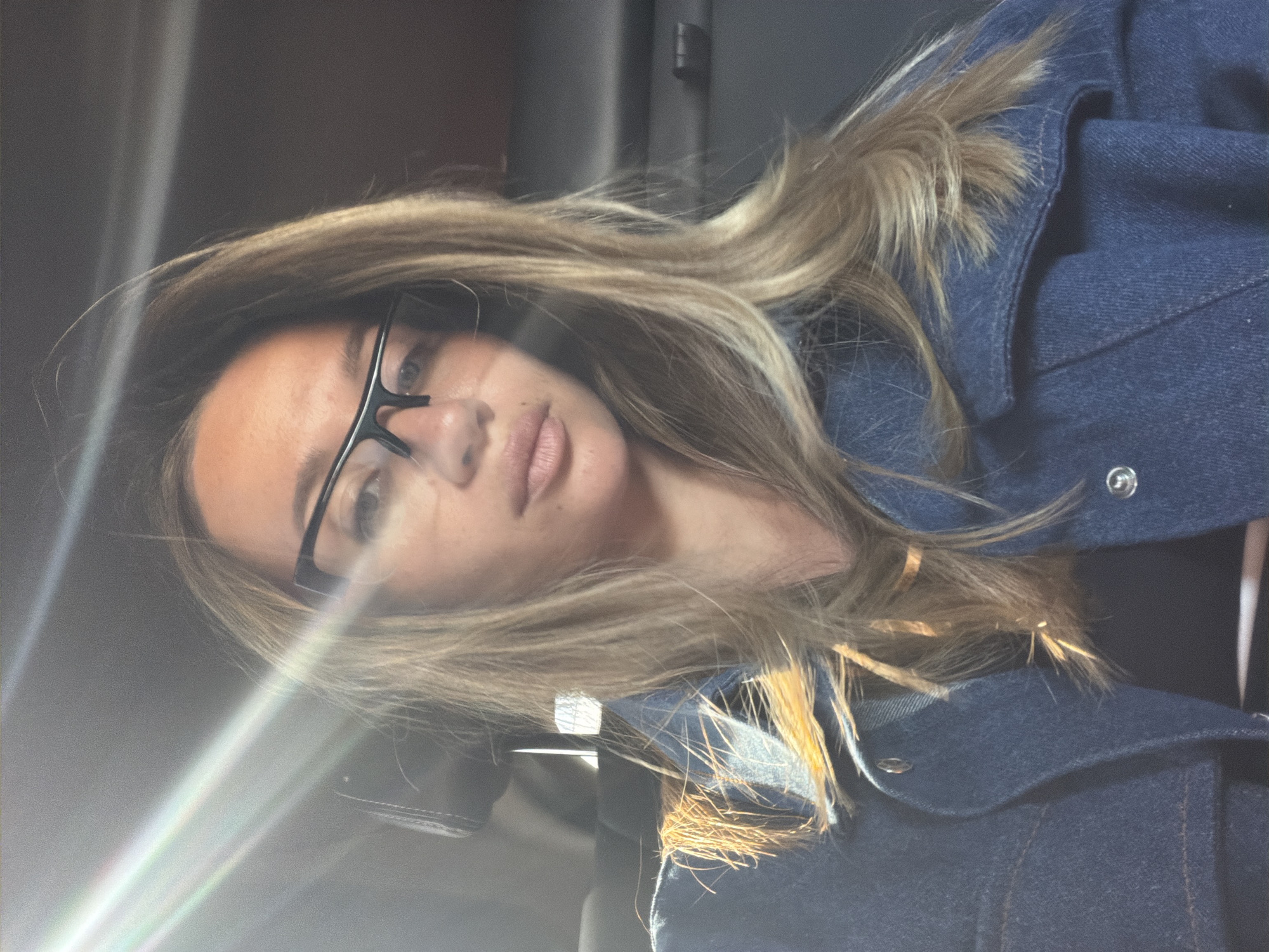 A woman with shoulder-length, blonde-highlighted hair and glasses looks directly at the camera. She is wearing a dark denim jacket. The background appears to be a dark interior, possibly a car, but details are obscured. A bright glare of light diagonally crosses the bottom left of the image. The location is unspecified.