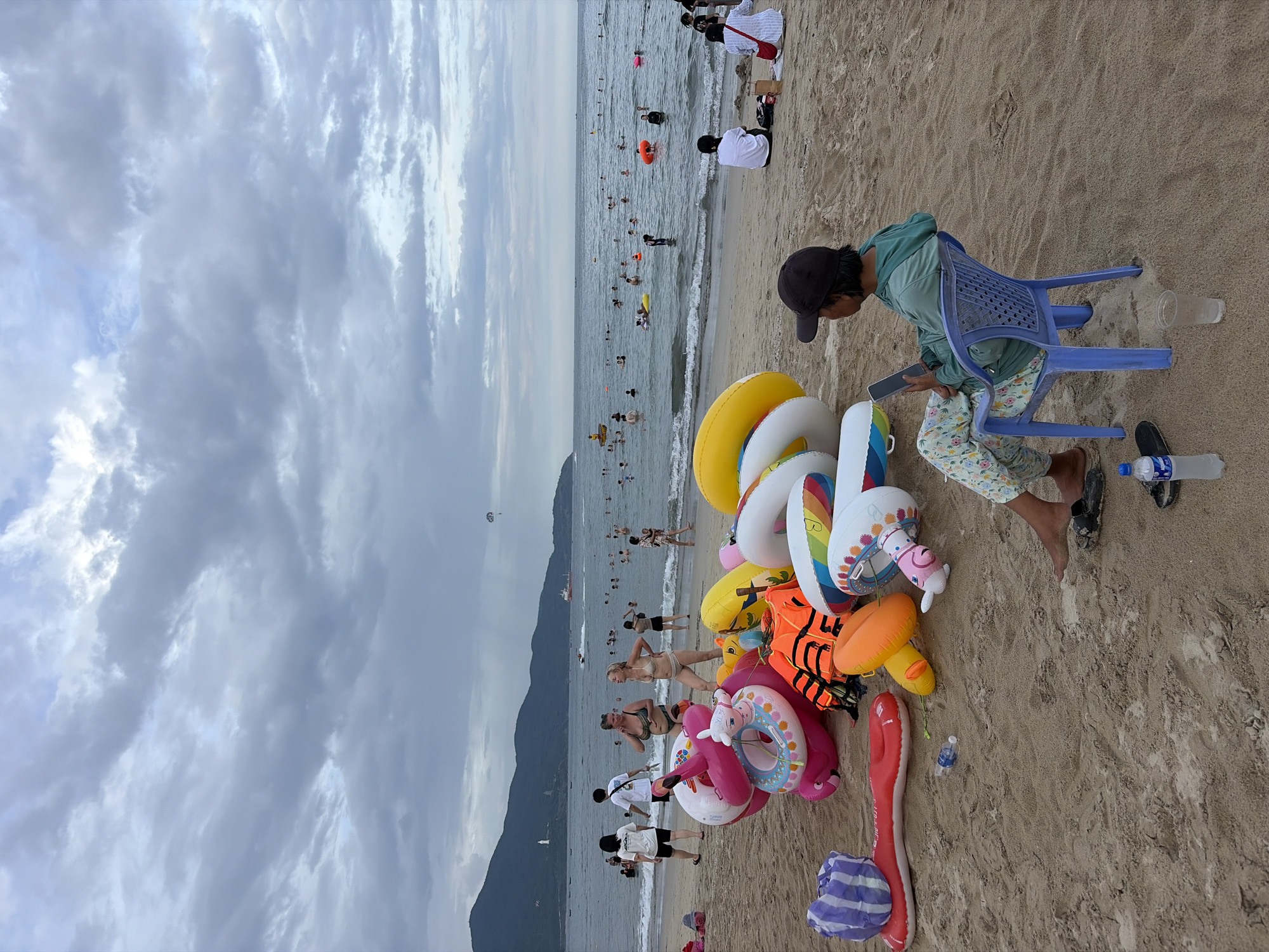 The image depicts a beach scene on a cloudy day, likely in Đà Nẵng, Vietnam, given the presence of a parasailer in the distance and the coastal mountain range. In the foreground, a collection of inflatable swimming toys, including a large pink flamingo, orange mushroom, and various rings, are piled on the sand. A person sits on a blue plastic chair, looking at a smartphone. In the middle ground, people are swimming and wading in the ocean. The background shows a coastline with mountains and a cloudy sky. There are also boats and a paraglider visible on the water. Two women in swimsuits are standing near the inflatable toys, facing away from the camera.