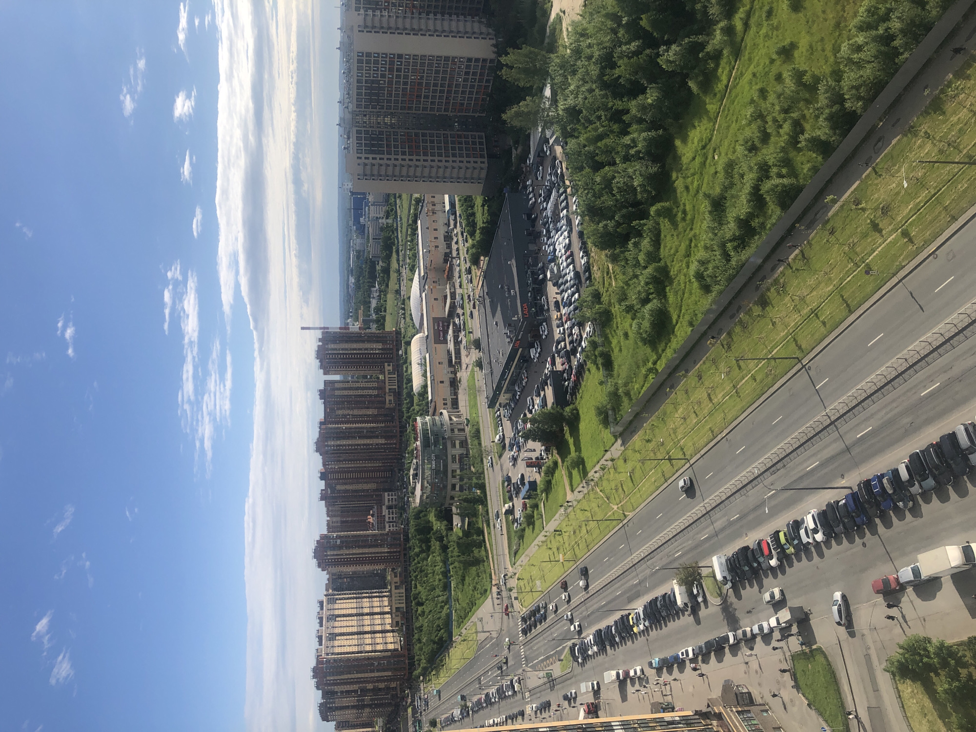 The image shows an aerial view of a city street on a sunny day, with tall residential buildings lining one side of the road and a grassy slope and trees on the other. The road is busy with traffic, with many cars parked along the side and others driving. A large parking lot filled with cars is visible near some of the buildings, possibly for a shopping center or residential complex. The sky is blue with scattered white clouds, suggesting a pleasant day. There is no visible text in the image.