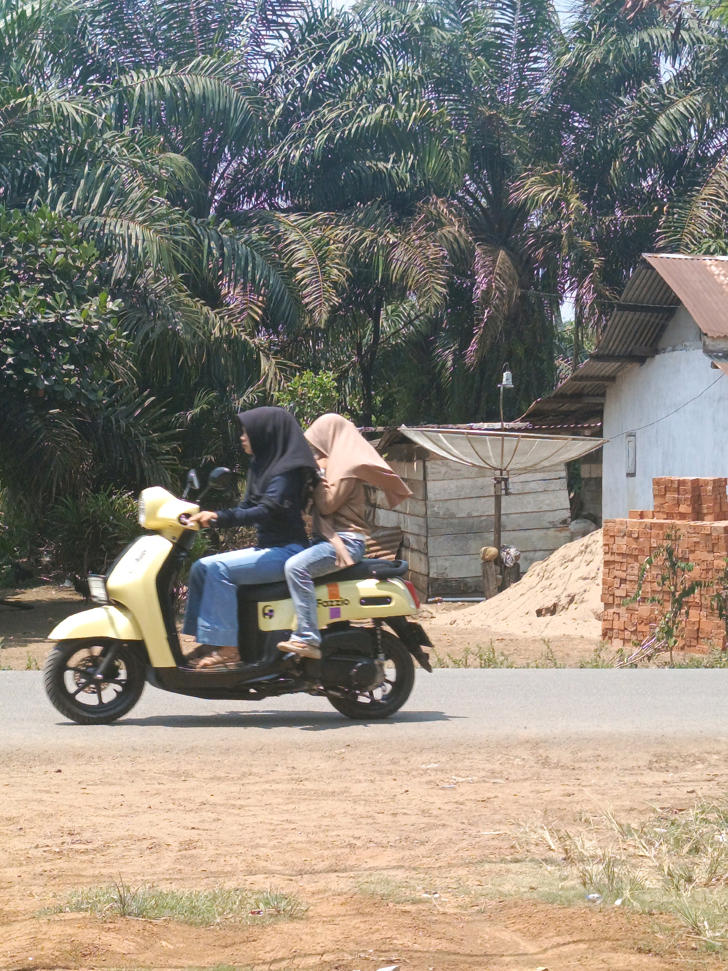 Two veiled women are riding a yellow scooter on a paved road in a rural area. The woman in front is wearing a black hijab and a dark blue top, while the woman in the back is wearing a light brown hijab and a brown top. They are both wearing blue jeans. The scooter has "FAZZIO" written on the side. Behind them, there are palm trees, a building with a corrugated metal roof, a satellite dish, a pile of sand, and a stack of bricks. The ground is covered in dirt and sparse vegetation. The scene appears to be in Megang Sakti I, Indonesia, based on the provided context. The lighting suggests it is daytime, likely a sunny day.