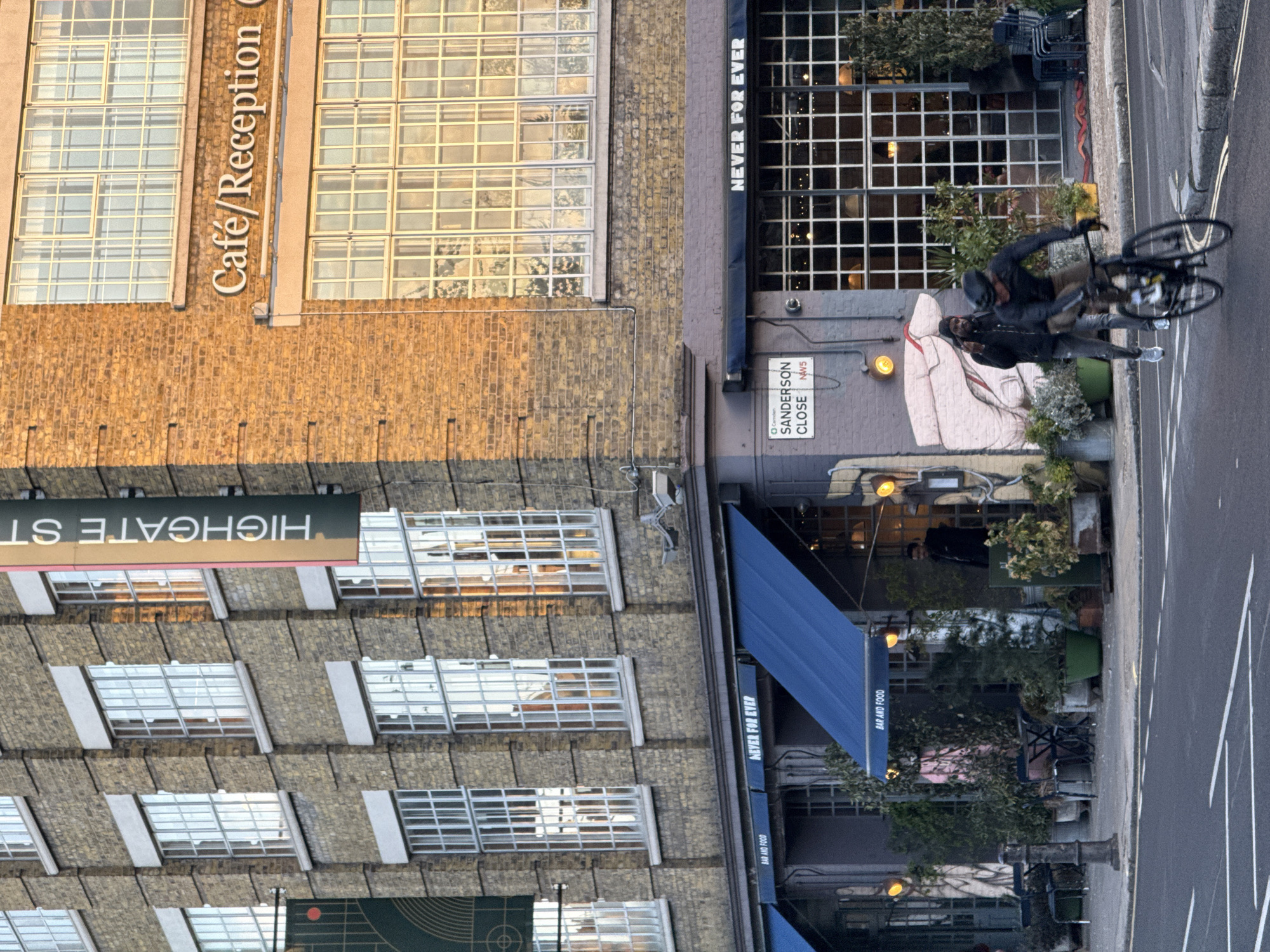 The image depicts a street scene in London, United Kingdom, specifically at Sanderson Close NW5, Camden. Several buildings are visible, including one with a sign that reads "Café/Reception" and another with a blue awning and the text "NEVER FOR EVER" and "BAR AND FOOD." Another sign is present, displaying "HIGHGATE ST." A mural is visible on a wall with a face on it. Two individuals are riding bicycles on the pavement. There are plants in pots, tables and chairs along the front of the building. A grey street with dashed white lines along with parked vehicles are also visible in the image.