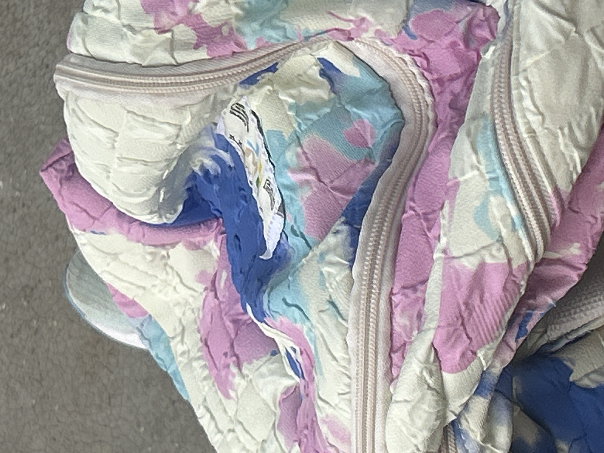 The image shows a close-up, overhead view of a folded, quilted fabric with a colorful, abstract pattern of pink, blue, and white. A zipper runs along the edge of the fabric. The fabric appears to be part of a garment or bedding. The background is a textured, gray surface, possibly asphalt or concrete, suggesting an outdoor urban environment. There is no indication of the specific location within Santa Ana, Venezuela.