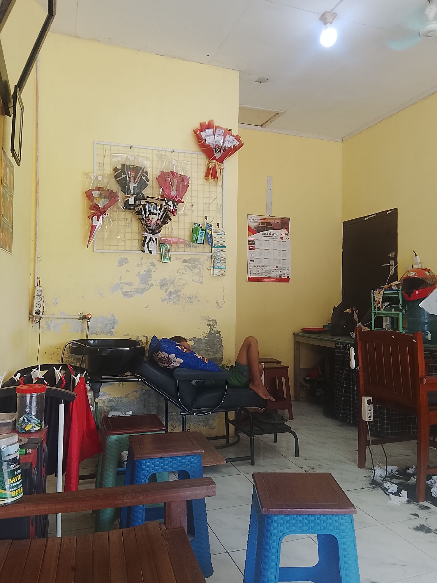 The image depicts an indoor scene, likely a barbershop or salon, in Karangklesem, Indonesia. A person is lying down on a reclining chair, covered with a patterned blue blanket, appearing to be asleep or resting. The room has pale yellow walls with visible signs of wear and tear.
On the wall, a grid display holds several decorative bouquets. A calendar with the year 2026 is also mounted on the wall. To the right, a dark wooden door is visible, and next to it, a green shelf unit holds various items, including a red helmet and what appears to be a large water bottle.
In the foreground, several small plastic stools are arranged. One is blue with a dark brown seat, another is green with a dark brown seat, and a wooden bench is also present. Various items are scattered around the floor and on surfaces, including plastic containers, bags, and what looks like discarded paper or fabric. A sink is positioned near the reclining chair. The lighting in the room comes from a single ceiling light fixture.