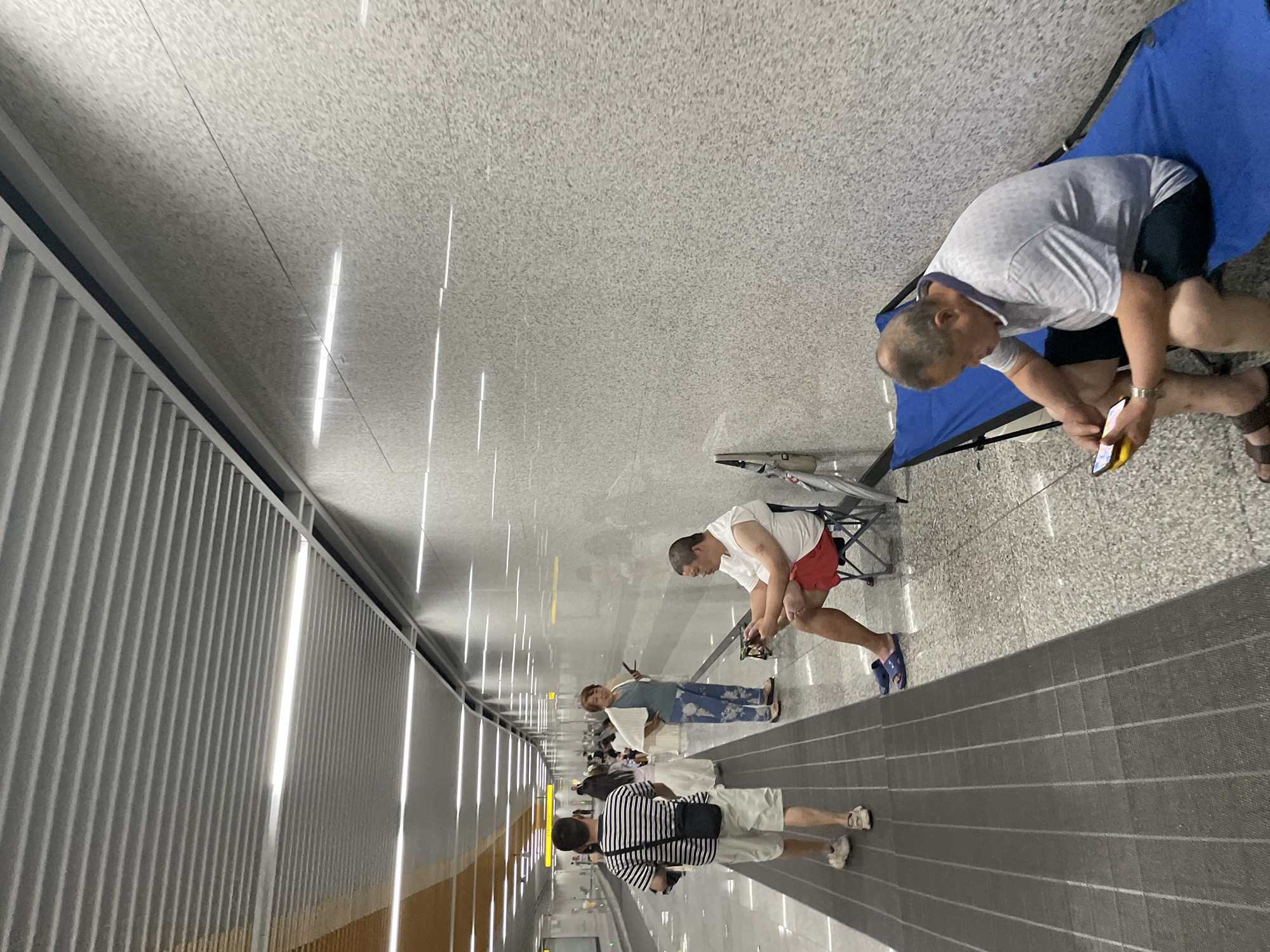 The image depicts a scene in a modern, indoor public space, likely a transportation hub, with a man sitting on a blue cot and looking at his phone. He is wearing a light gray patterned t-shirt, dark shorts, and sandals. To his left, another man is sitting in a folding chair, wearing a white t-shirt, red shorts, and blue slip-on shoes, and appears to be looking at a handheld device. Further down the hallway, several other people are standing and walking, including a woman in a green top and patterned pants holding a phone, and a man in a striped t-shirt and shorts walking away from the camera. The environment features a textured gray wall, a patterned floor, and a suspended ceiling with integrated lighting. The image was taken in the city of Liangjiang, China.