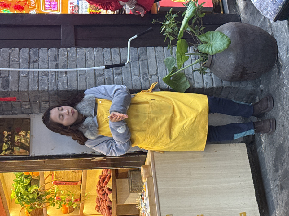 A woman in a yellow apron and blue jacket stands outside a shop, eyes closed, hands clasped. Behind her, a gray brick wall with a white bar extends from the wall. A large, dark pot sits beside the wall with a green plant growing out of it. Shop items, including oranges and sausages, are visible on the left. The woman wears dark pants with blue stripes at the bottom, and brown shoes.