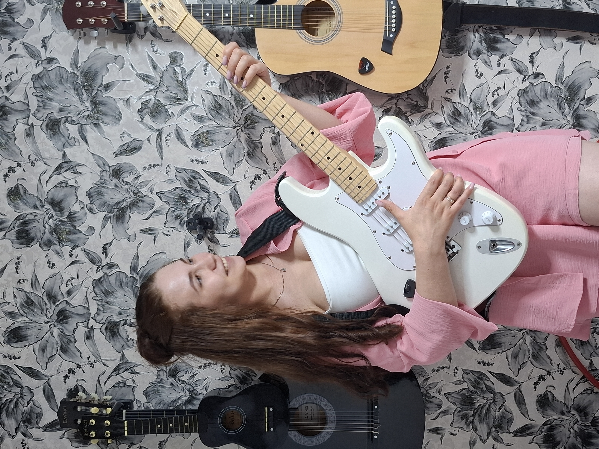 A woman lies on a floral-patterned surface in what appears to be a home setting, possibly in Chaikovsky, Russia. She wears a white top under a pink, long-sleeved shirt or jacket and shorts, and has brown hair styled in a bun. Around her neck is a delicate necklace. She is holding and looking at a white electric guitar, which is slung over her shoulder with a black strap. Above and below her are acoustic guitars - one light brown and one black. A guitar pick is stuck on the light brown guitar. A red cord is visible on the right edge of the frame.