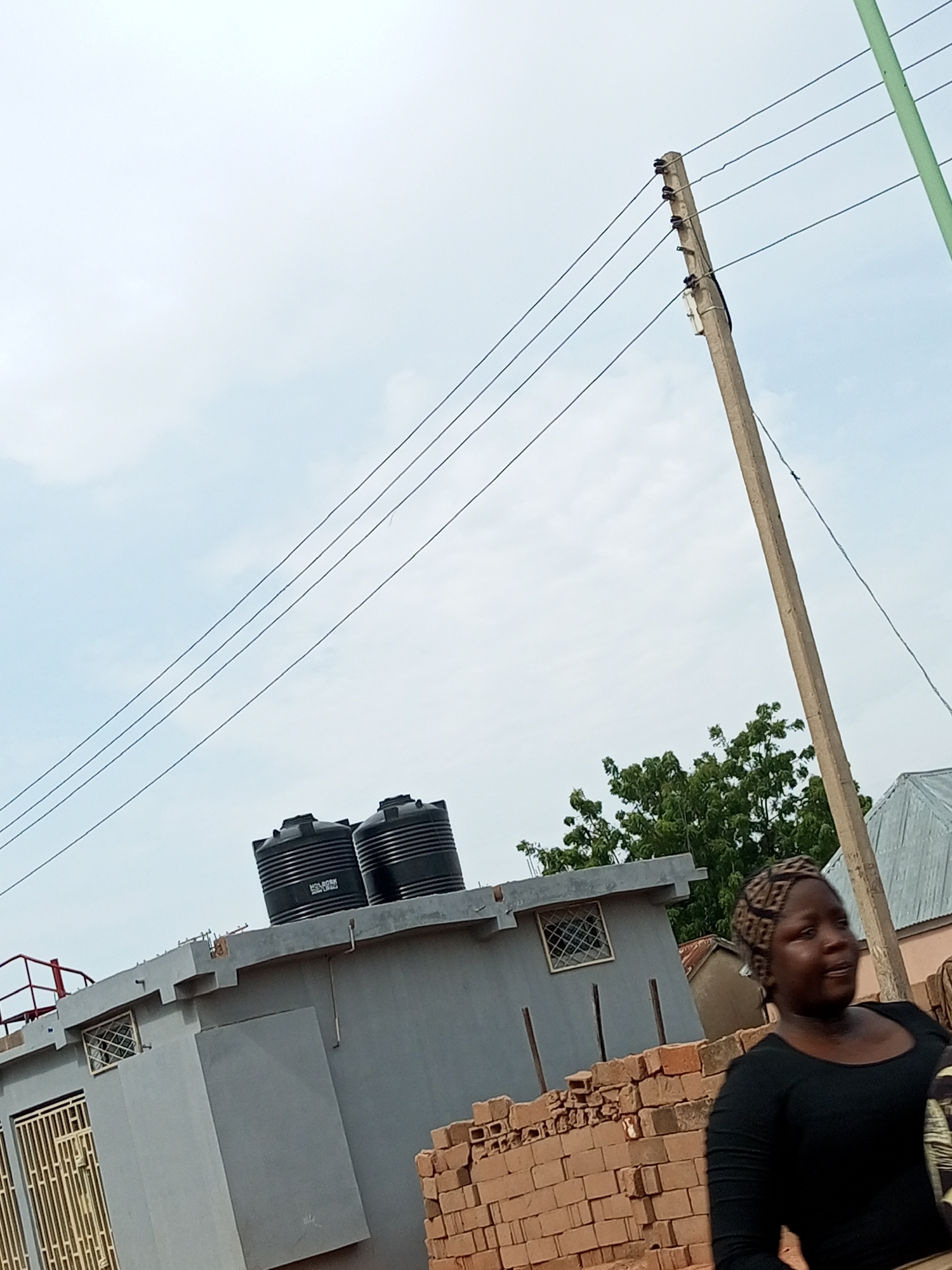 In Tumfure, Nigeria, a person wearing a black shirt and a patterned headscarf stands in the foreground, facing to the right. Behind them, a stack of bricks is visible. Further back, a grey concrete structure with two large black water tanks on its roof is situated. Overhead, power lines stretch across the sky, supported by a wooden utility pole. Green trees and parts of buildings with corrugated metal roofs can be seen in the background. The scene appears to be outdoors, under an overcast sky.