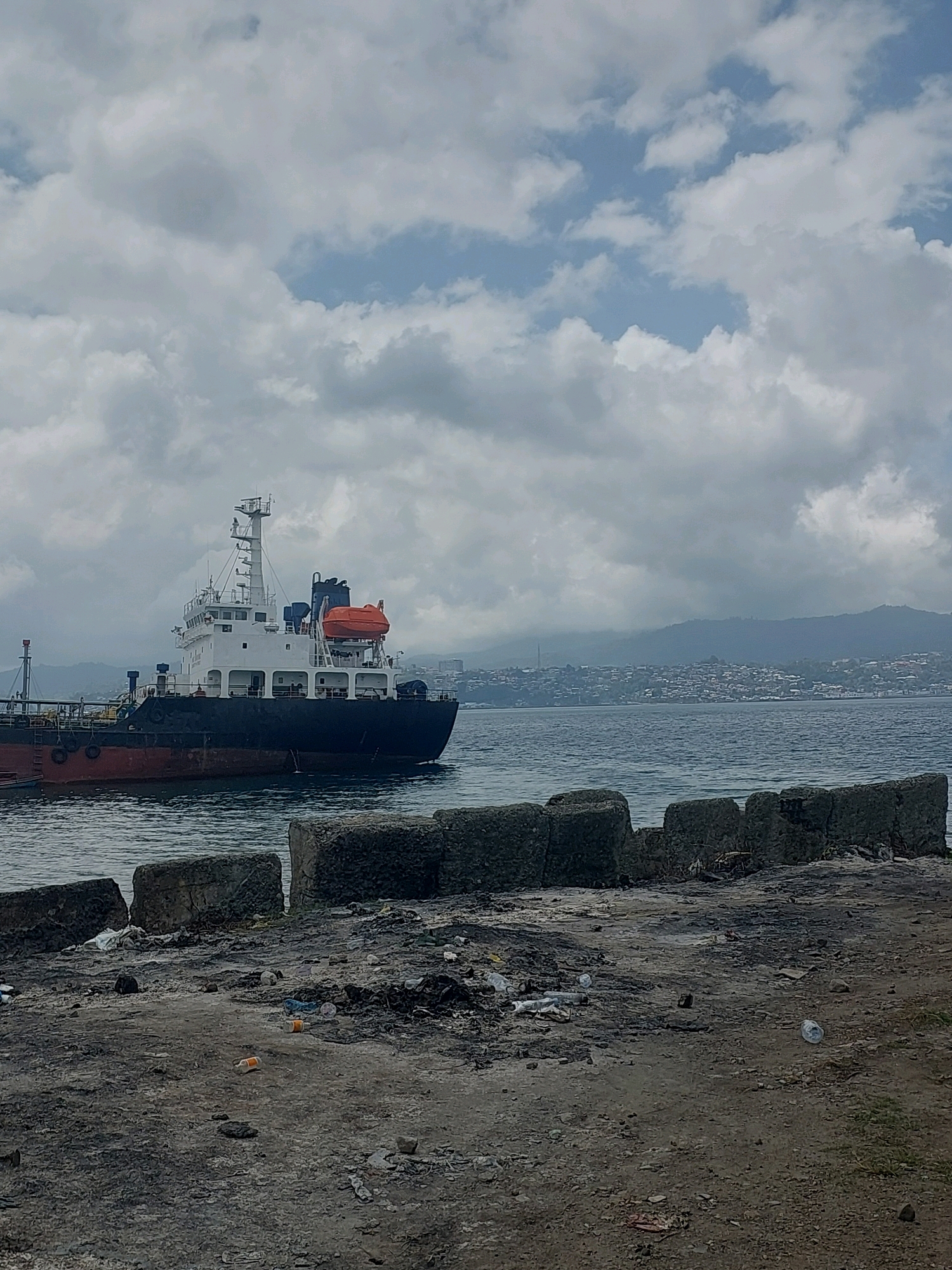 The image depicts a large cargo ship docked near a city on a body of water. The foreground shows a cluttered, unkempt shoreline with concrete blocks and debris scattered across the ground. The water is calm, and the sky is overcast with scattered clouds. In the background, a densely populated city is visible on a hillside, suggesting an urban coastal environment. The ship itself is white and dark, with visible railings and a prominent orange life raft. The city in the distance appears to have many buildings clustered together, with some mountainous terrain behind it. The overall impression is one of a working port area with signs of industrial activity and a nearby urban center. This scene is consistent with a view of the Ambon Bay area in Ambon, Indonesia, known for its port facilities and the characteristic hilly topography of the city.