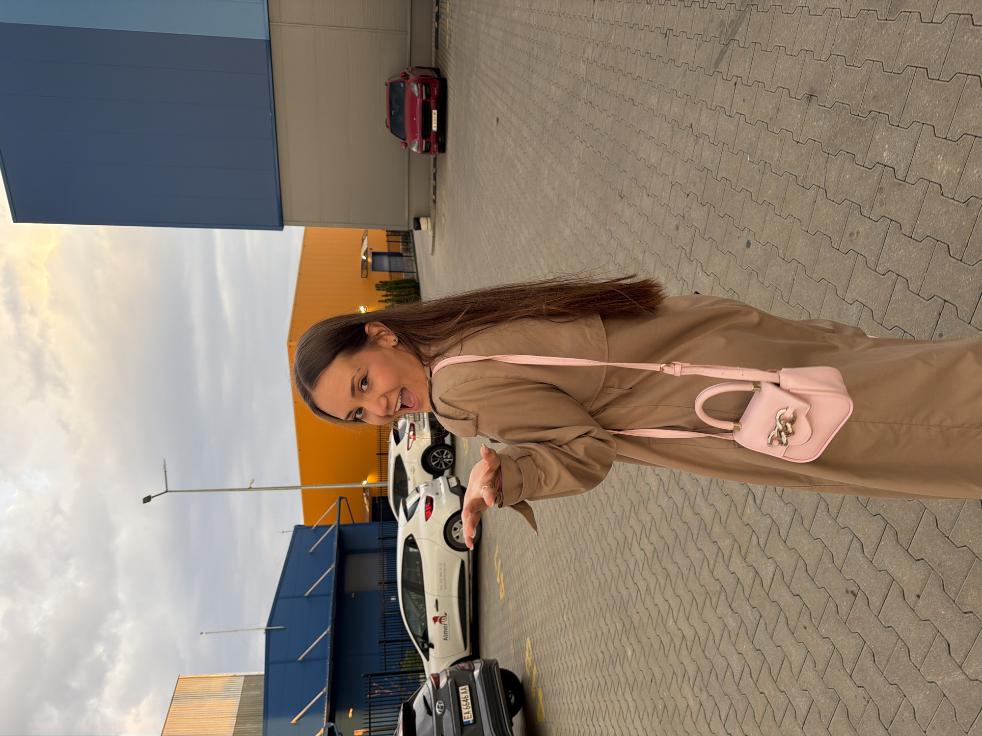 A young woman with long, straight brown hair is depicted smiling joyfully and making a playful gesture with her right hand. She is wearing a light brown trench coat and carrying a small, light pink purse with gold chain embellishments over her left shoulder. Her expression is bright and animated, with her mouth open in a wide smile.
The scene takes place outdoors, likely in a large parking lot or industrial complex in Sofia, Bulgaria, as indicated by visible textual cues. The ground is paved with grey interlocking concrete blocks. In the background, several cars are parked, including a red car further back, and closer to the woman, a white car and a dark grey car. The white car features text on its side, reading "Tel: 0700 444 30" and "Aimer Rental Cars." The dark grey car has a license plate "EA 6846 AA," consistent with Bulgarian plates, with "EA" denoting the Sofia region.
Behind the vehicles, large, unadorned industrial or commercial buildings are visible, one in a distinct dark blue color and another in yellow or orange. The sky above is overcast with clouds, suggesting daytime, possibly late afternoon. A tall street lamp stands against the sky. The overall impression is candid and cheerful.