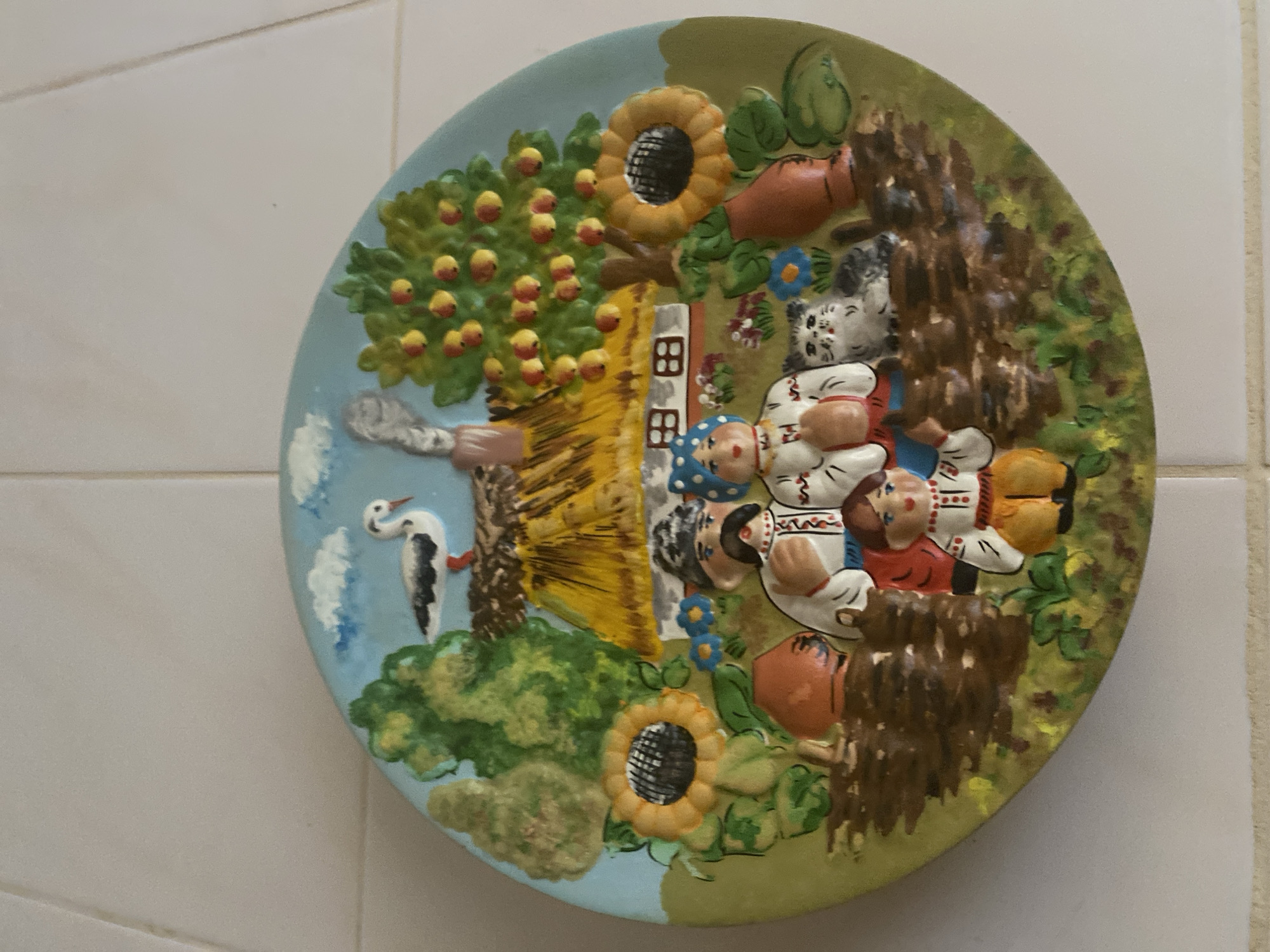 A circular, decorative plate with a relief scene depicts a family in front of a thatched-roof house. The scene is set in a rural environment with trees, flowers, and a bird. The family appears to be composed of two adults and two children. The adults are dressed in traditional clothing, with the man wearing a hat and the woman a headscarf. The children are also in traditional attire. A cat sits to the right of the family. The house has a smoking chimney. The plate is positioned on a tiled surface, likely a wall.