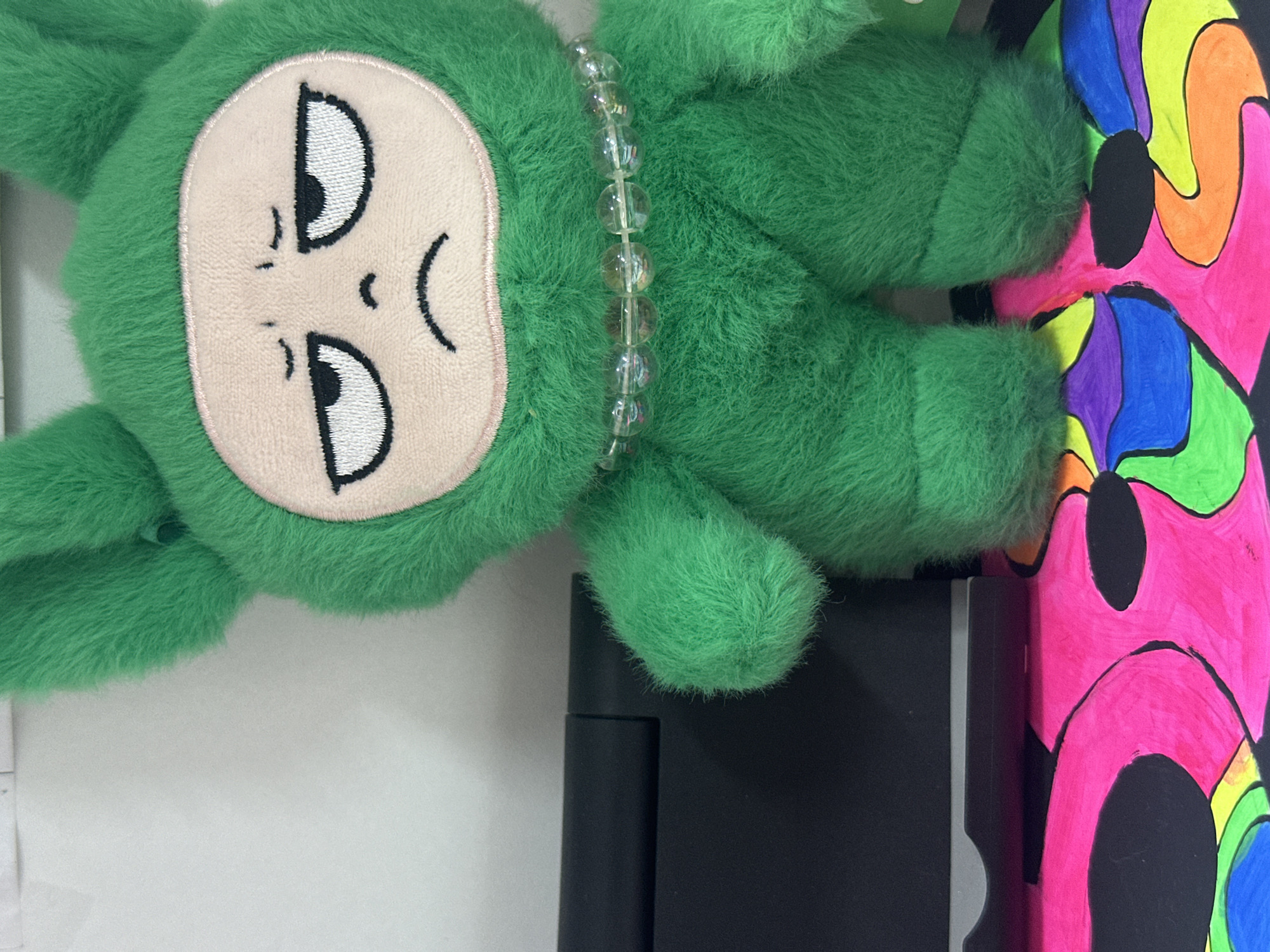 A green stuffed toy with a cartoon face, embellished with a clear beaded necklace, lies on top of a closed, black laptop and a colorful, abstract painting. The background is a neutral-toned surface.