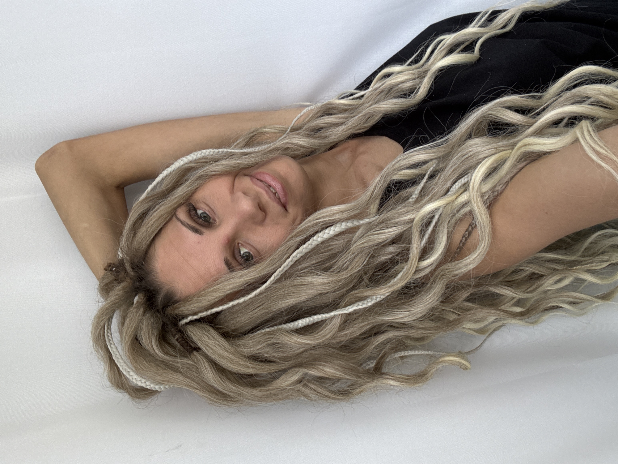 A woman with long, light brown and blonde hair that includes thin, white braids is lying down on a white surface. She is wearing a black top. She is looking at the camera with a slight smile. Her left arm is raised and bent, resting near her head. There is no evidence to indicate that this photo was taken in Rusanovo, Russia.