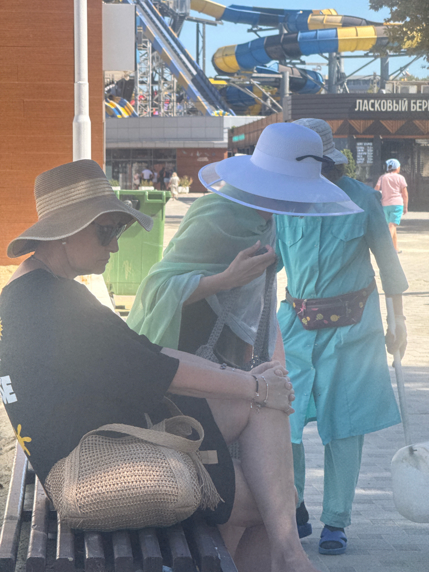 In Anapa, Russia, at what appears to be a water park, three individuals are visible. On the left, a woman wearing a wide-brimmed straw hat and sunglasses is seated on a wooden bench. She is dressed in a dark t-shirt and has a woven straw bag at her side. In the center, another person is partially obscured, wearing a light green scarf or shawl and a white wide-brimmed hat with a clear plastic brim. To their right, a third individual, possibly male, is wearing a light blue tunic and pants, a fanny pack with a pineapple pattern around their waist, and blue sandals. They are holding a white walking stick. In the background, a large water slide structure with blue and yellow tubes is visible, along with buildings and other park-goers. The ground is paved with light-colored stones.