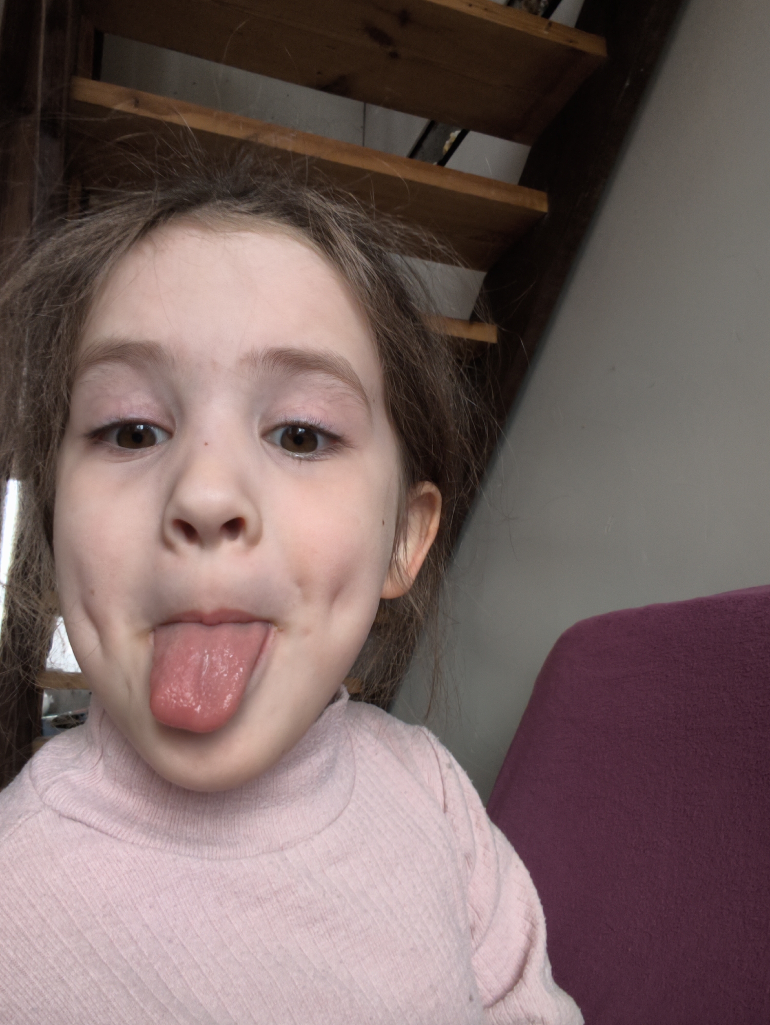 A young girl is sticking her tongue out at the camera. She is wearing a pink turtleneck shirt. Her hair is brown and slightly messy. Behind her, there is a wooden staircase and a grey wall. Part of a purple cushion or piece of furniture is visible on the right. The image does not contain enough information to determine its location within the city of Прикамский, Russia.
