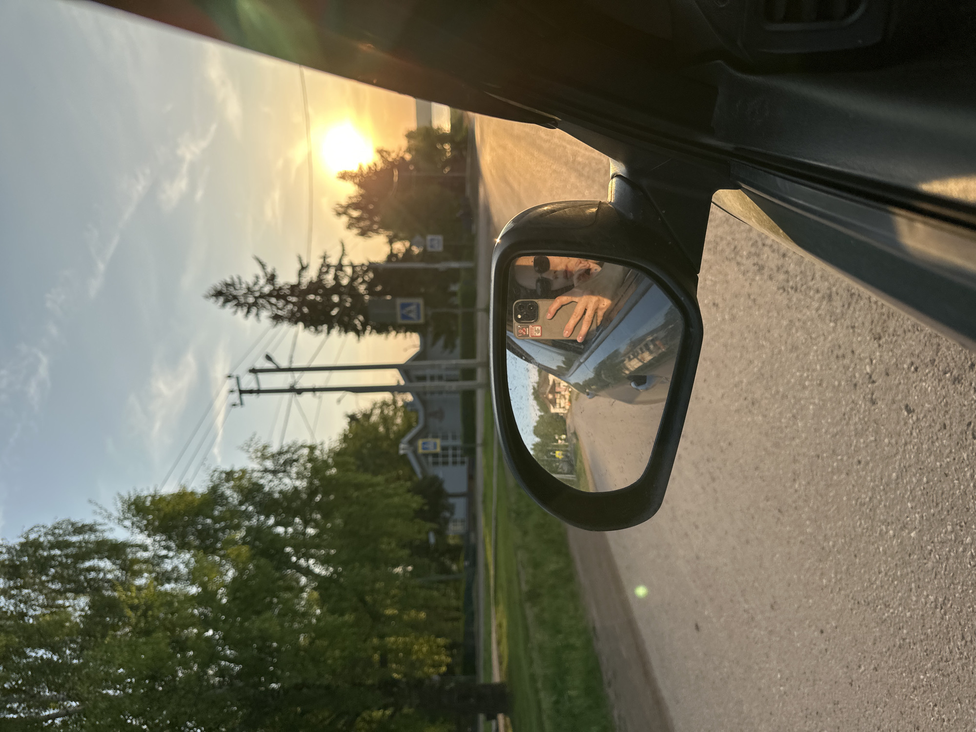 The image depicts the view from inside a car, looking through the driver's side window and side mirror. The sun is setting in the distance, illuminating the road and trees. Reflected in the side mirror is a person taking a photo of themselves with a smartphone. Outside the car, there are houses, trees, power lines, and a pedestrian crossing sign. The location is identified as Ilyinsky, Russia.