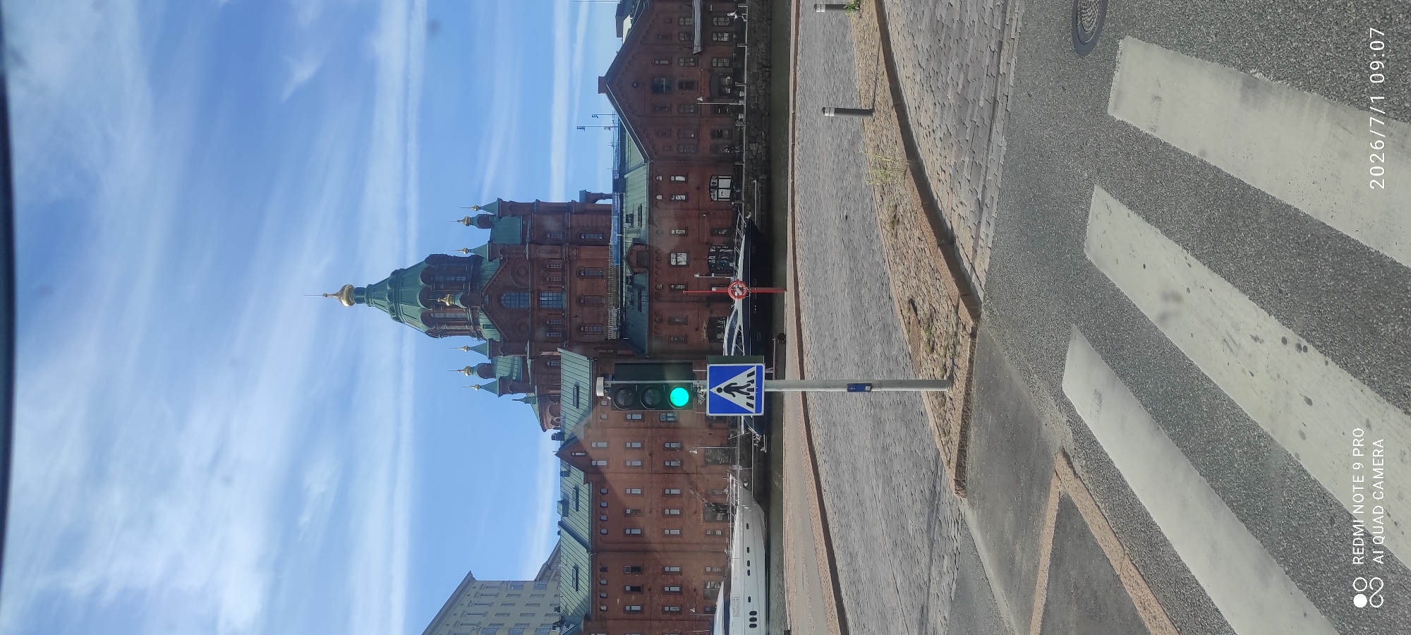 In a city scene, a pedestrian crossing is in the foreground with a traffic light showing green. A sign for a pedestrian crossing is also visible. Behind this, a large brick building with a green roof and a prominent domed structure topped with gold is situated. Several boats are docked in the water in front of the building, and the sky is blue with scattered clouds. The overall environment appears to be urban and waterfront.