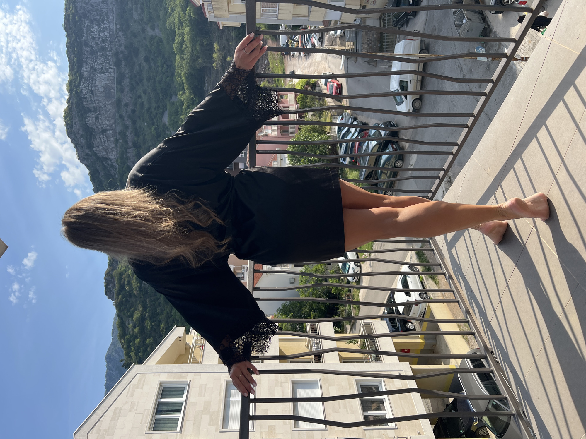 The image captures a woman from behind, standing on a balcony in Budva, Montenegro, and leaning on a dark metal railing. She has long, light brown hair and is wearing a black robe or short dress with distinctive black lace trim on the cuffs of its wide sleeves. Her bare feet are visible on the sunlit, tiled balcony floor.
The balcony offers a view of a street below, where several cars are parked. Across the street, other multi-story residential buildings with windows and balconies can be seen. In the background, a prominent, steep mountain rises, displaying a mix of rocky surfaces and green foliage under a clear blue sky dotted with a few scattered white clouds.
The strong shadows cast by the railing on the balcony floor indicate bright sunlight, suggesting it is late morning or early afternoon. The scene conveys a sense of tranquility as the woman gazes out at the urban landscape and the natural beauty of the mountain and sky. No text is visible in the image.