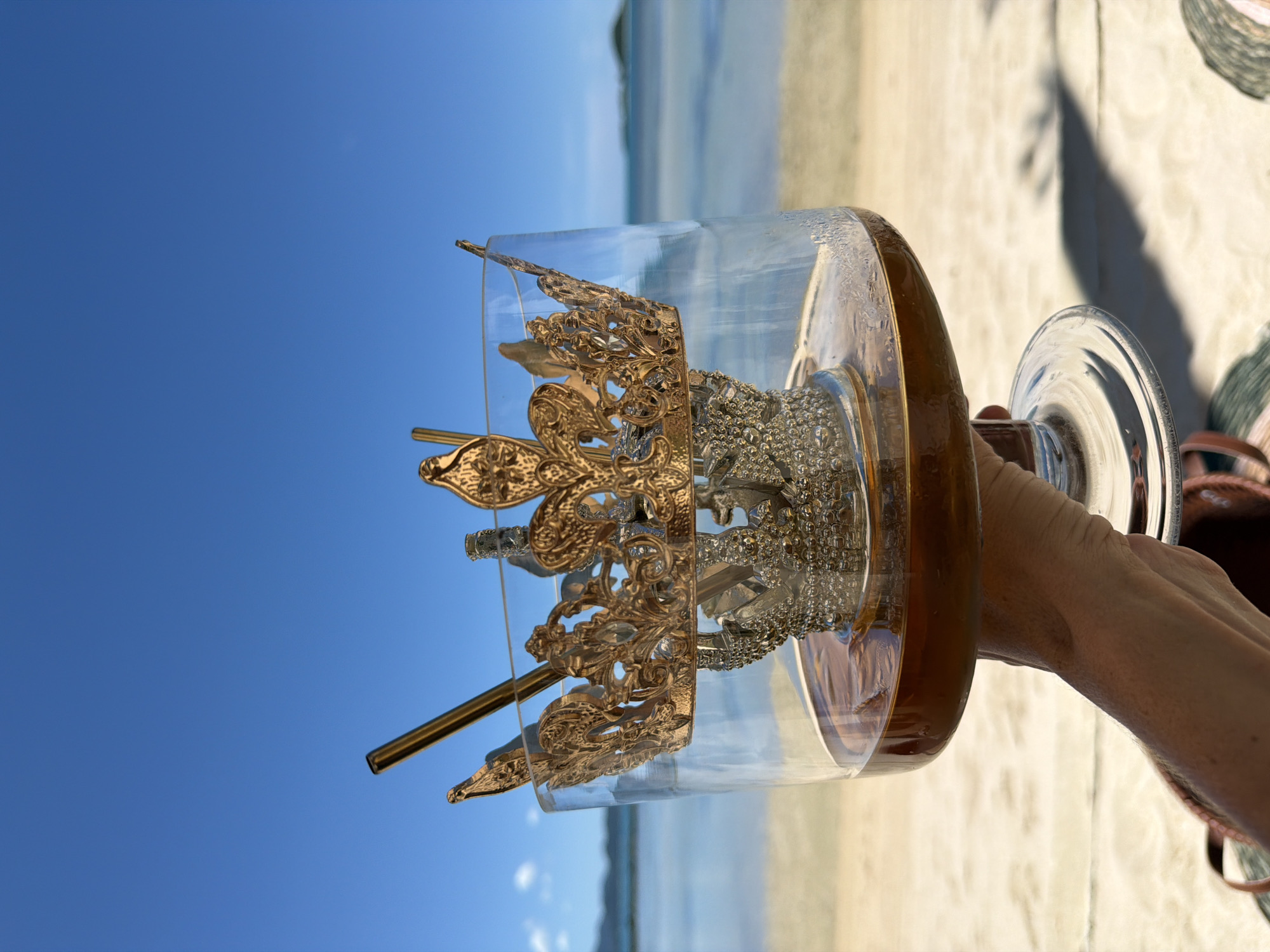 A person's hand holds a clear glass goblet filled with a dark amber liquid and two gold straws. A crown, decorated with gold filigree and silver embellishments, sits inside the goblet. The background features a beach with sand, calm ocean water, and a clear blue sky with a few white clouds. The scene is set in Baan Thong Krut, Thailand.
