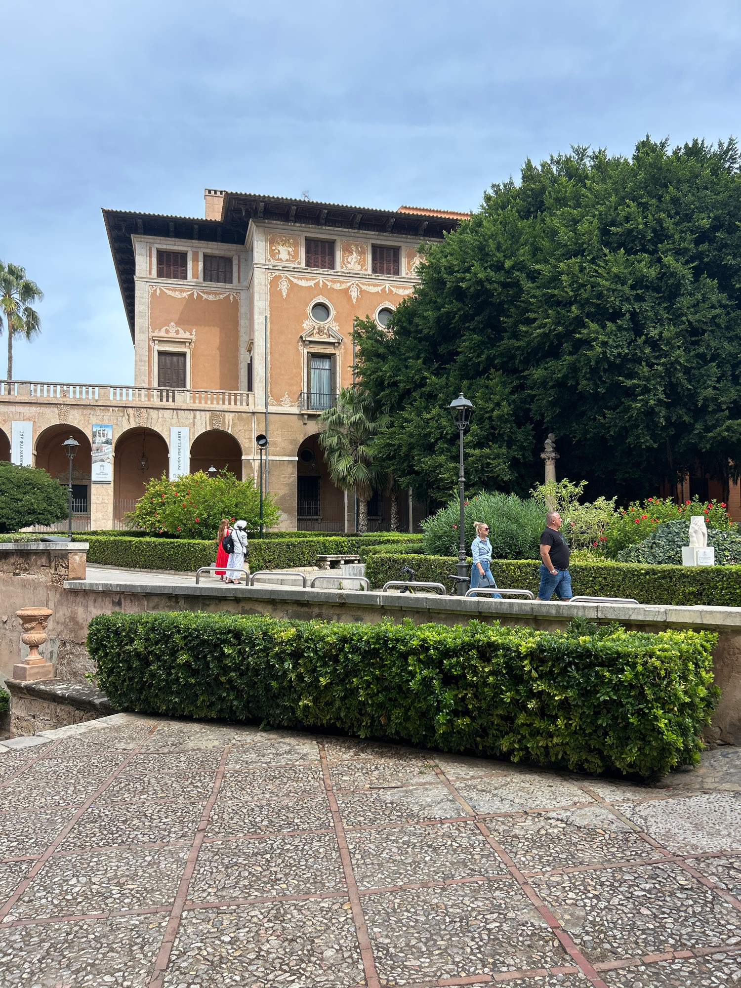 This image depicts the Palau de l'Almudaina in Palma de Mallorca, Spain. The foreground shows a paved walkway made of small, irregular stones interspersed with rectangular brick borders, leading up to a manicured hedge. Behind the hedge, a raised walkway is visible, where two individuals are walking. One person is wearing a white outfit, and the other is in a red dress. Further back, several more people are seen walking along another elevated pathway in front of a large, ornate building. The building features a distinctive architectural style with arched colonnades, numerous windows, and decorative ornamentation on its facade. A lush, dense green tree dominates the right side of the frame, and palm trees are visible on the left, suggesting a Mediterranean climate. The sky is overcast, creating a soft, diffused light.
