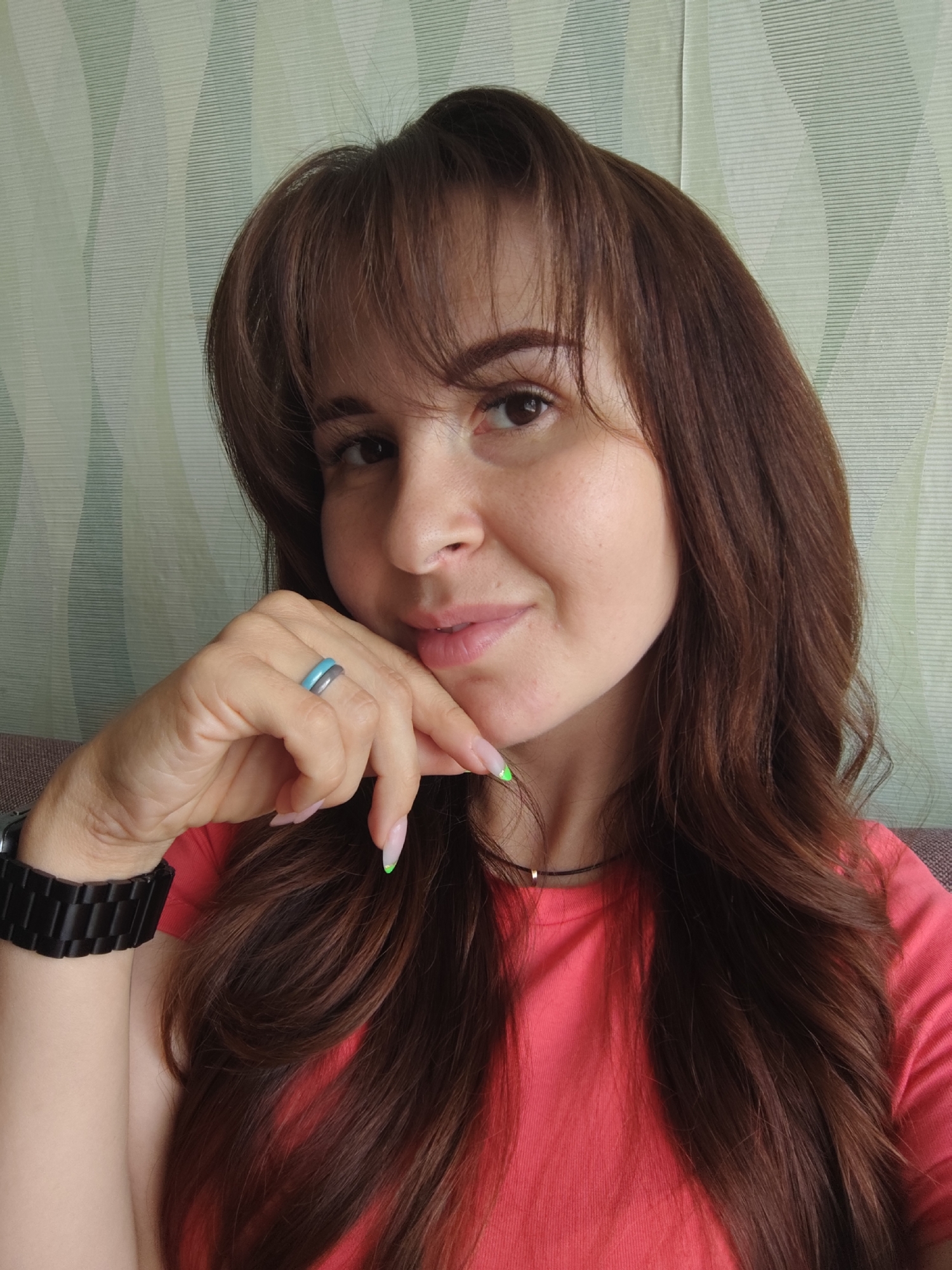 A woman with long, brown hair and bangs is shown from the chest up, looking at the camera with a slight smile. She is wearing a coral pink t-shirt and a black watch on her left wrist. Her right hand is raised to her face, with two rings on her fingers. Her fingernails are painted with a light pink polish, with the tips of two nails featuring a neon green accent. The background is a textured wallpaper with a pattern of vertical wavy lines in varying shades of green and white. The image appears to be taken indoors, possibly in a home environment. There is no indication of the location being in the city of Fokino, Russia.