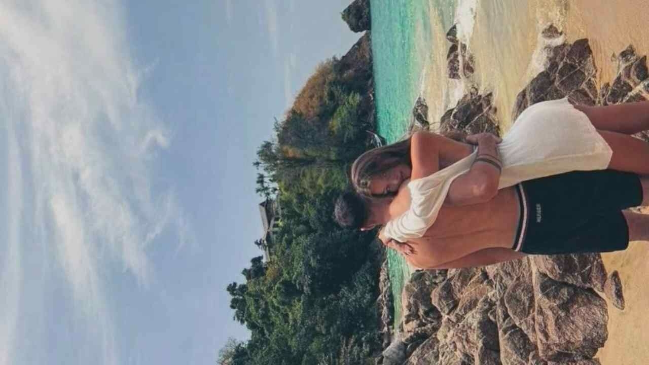 A young couple embraces on a rocky beach, their backs to the camera. The man, shirtless and wearing black swim trunks, is holding the woman from behind. The woman is wearing a white dress and has her arms wrapped around his neck. The ocean is visible behind them, with a lush, green hill dotted with buildings in the background. The sky is clear with some scattered clouds. The location is unknown, city unknown.