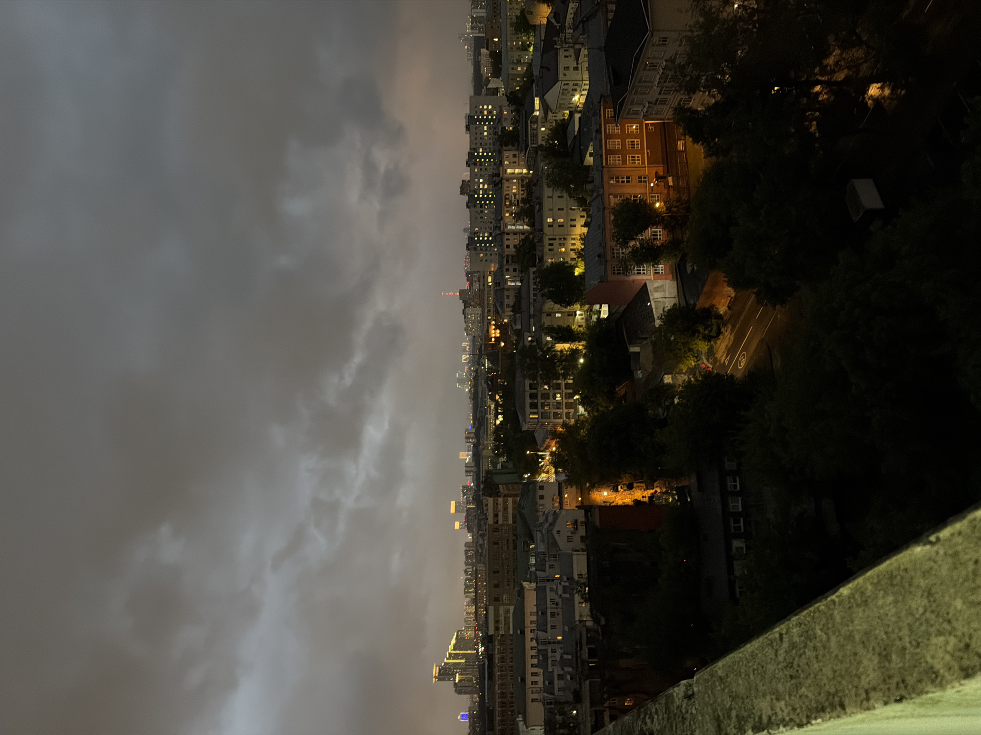 This is a nighttime aerial view of a cityscape in Moscow, Russia, likely taken from a high vantage point. The sky is overcast with dark, moody clouds, hinting at impending or ongoing inclement weather. Below, a dense urban environment unfolds, characterized by mid-rise apartment buildings and some taller structures in the distance. Many windows are illuminated, casting a warm glow onto the dark streets and facades. Trees are interspersed throughout the urban landscape, their foliage appearing dark and dense against the artificial lights. A concrete barrier or ledge is visible in the foreground, suggesting the photograph was taken from a balcony, rooftop, or similar elevated structure. The overall atmosphere is somber and quiet, typical of a city at night under a cloudy sky.