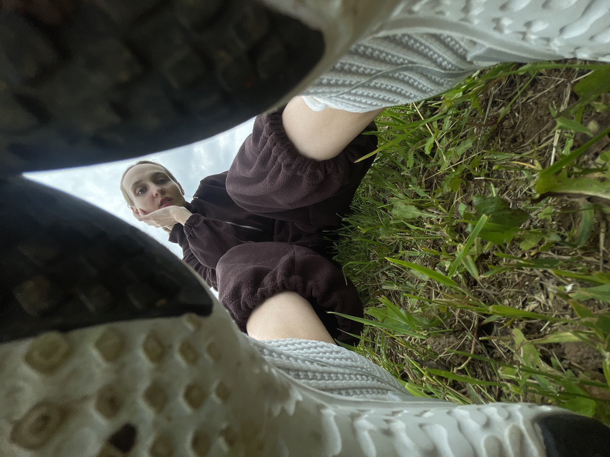 A low-angle shot captures a young Caucasian woman sitting on the ground in a grassy field. She is wearing a brown fleece tracksuit and white socks. Her legs are bent, and her feet are clad in athletic shoes, the soles of which are prominent in the foreground, framing the woman. Her gaze is directed towards the camera, with her right hand resting on her cheek. The background consists of green grass, dirt, and a cloudy sky. The location is specified as Podshivalovo, Russia.
