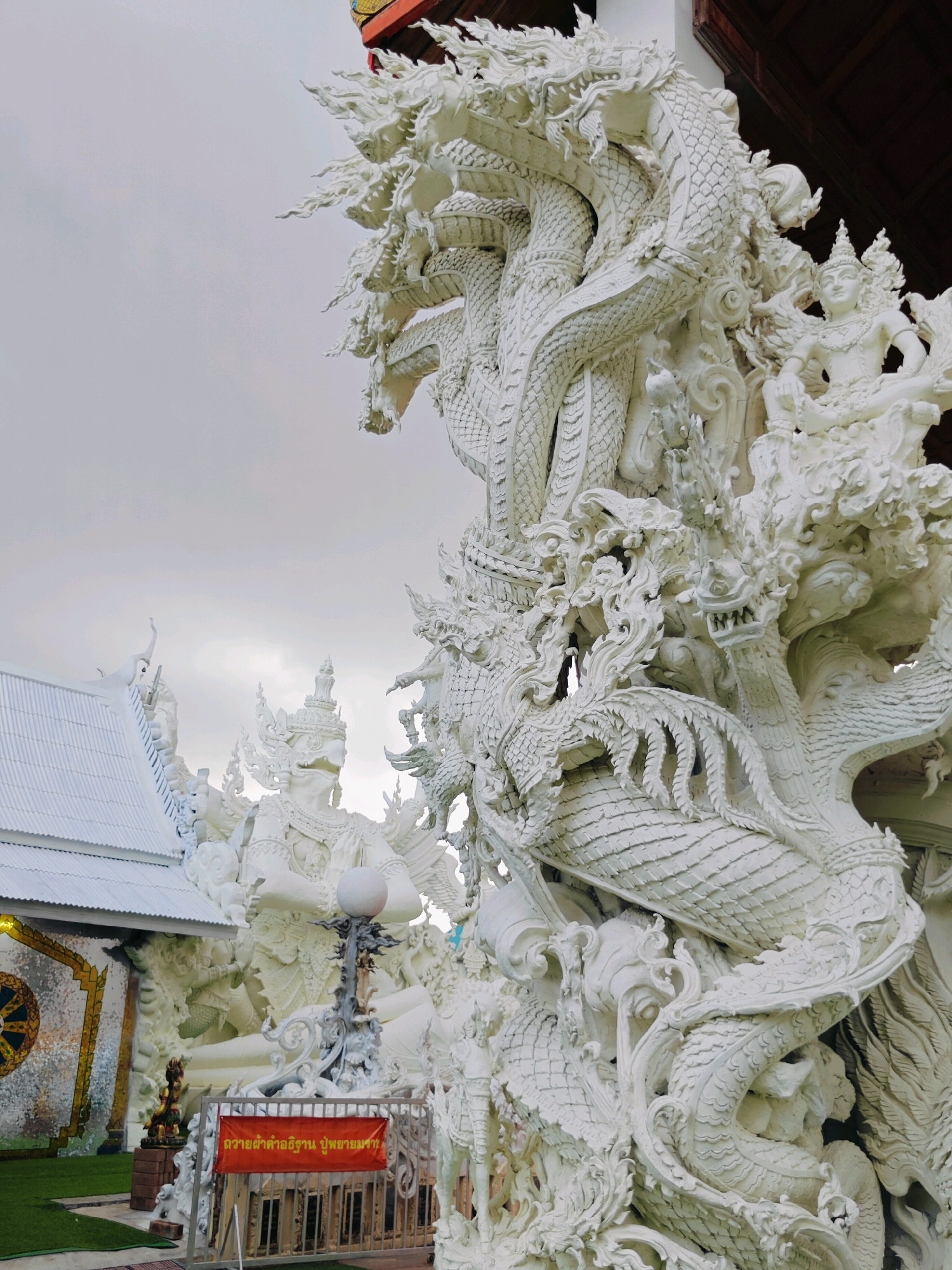 The image shows an intricate white stucco sculpture of a multi-headed serpent, likely a Naga, a mythological creature prominent in Southeast Asian cultures. The serpent is entwined with other mythical figures and decorative elements, creating a dynamic and detailed composition. In the background, a white temple structure with a distinctive tiled roof and a large circular mosaic is visible. The foreground features a metal fence with an orange banner containing Thai writing. The overall scene is characterized by its ornate white architecture and sculptures, suggesting it is a religious or cultural site. Based on the architectural style and the cultural references, this location is likely within Thailand, possibly in a historically significant area such as Phra Nakhon Si Ayutthaya City Municipality, known for its temples and historical ruins. The sky is overcast, creating a diffused light that highlights the texture of the stucco.