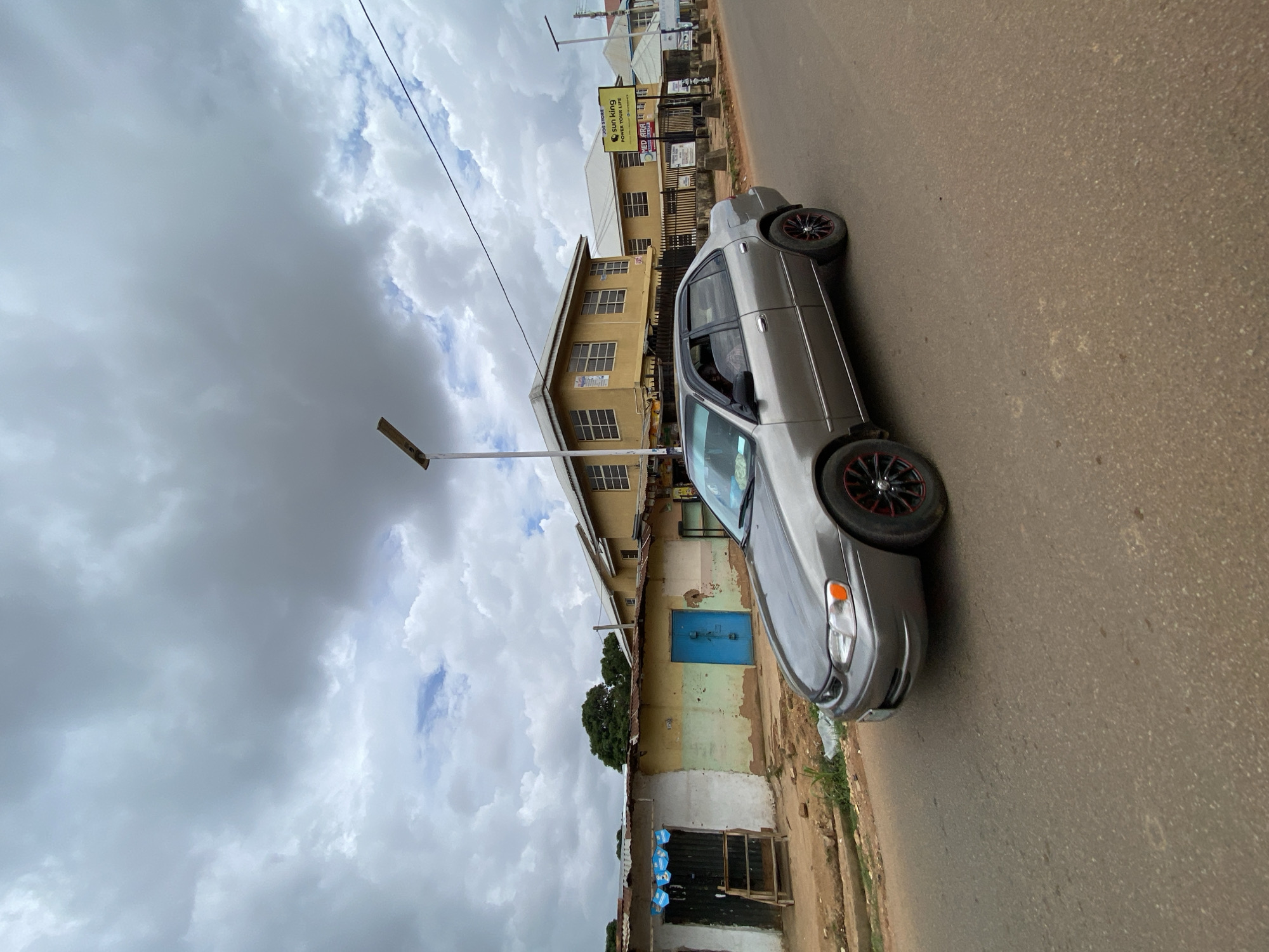A grey sedan is parked on a dirt road in Gura Top, Nigeria. The car has black rims with red accents. In the background, there are two yellow buildings with windows and a metal roof on a smaller structure. The sky is cloudy and overcast.
