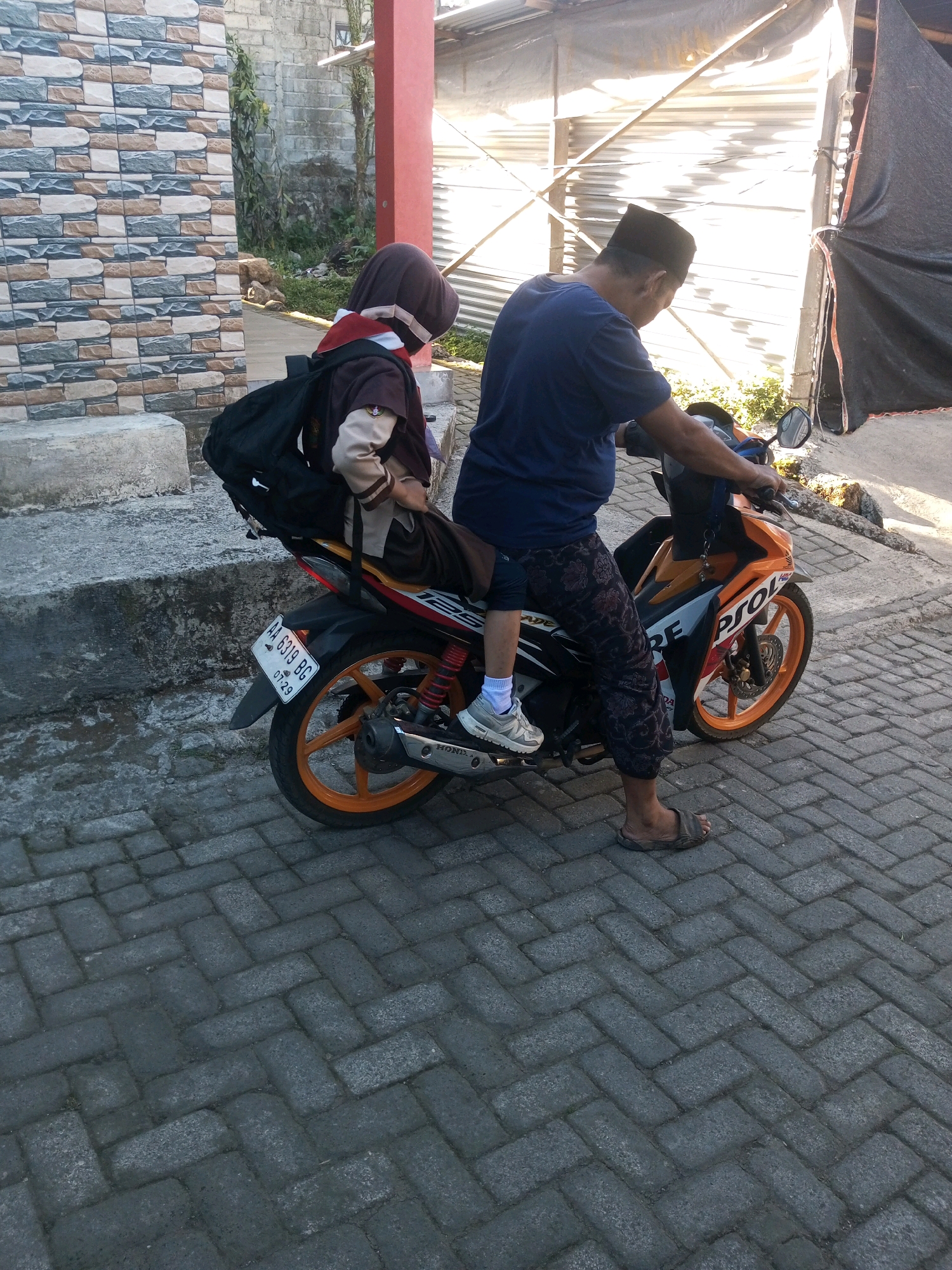 In Andongsili, Indonesia, on a paved brick sidewalk, a man wearing a blue t-shirt, patterned pants, and sandals is riding a motorcycle. He is wearing a black skullcap. A young person, wearing a brown and beige uniform with a red scarf, is riding on the back of the motorcycle. The young person has a black backpack on their back and is wearing white socks and gray sneakers. The motorcycle is orange and white with a license plate that reads "AA 6319 BG". In the background, there is a building with a tiled wall and a red pillar, and some vegetation. The scene appears to be during the daytime, with bright sunlight.