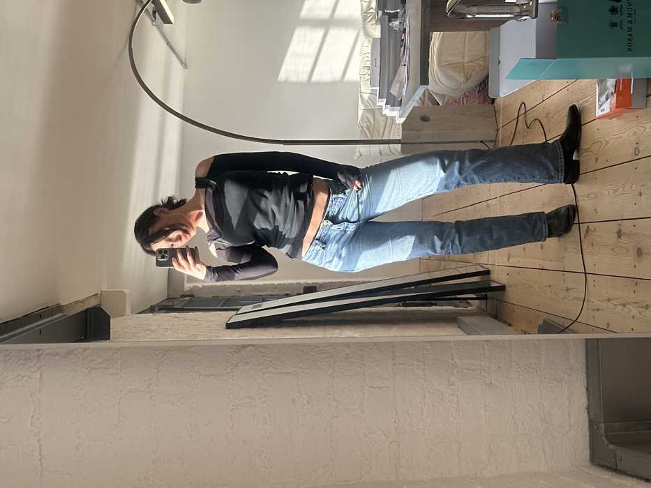The image depicts a female individual, approximately 20-35 years of age, capturing a mirror selfie within an indoor setting. She stands on light-toned wooden floorboards, dressed in a dark, long-sleeved top with cut-out shoulders, light blue straight-leg jeans with a button fly, and dark ankle boots. Her right hand holds a dark-cased smartphone towards the mirror, with her left hand resting near her jeans' front button. Direct sunlight from an unseen window to the right illuminates her and casts defined shadows on the floor and right wall.
The reflective background reveals a white brick wall to the left and a plain white wall to the right. Two narrow, dark-framed mirrors lean against the left wall. An arched dark metal floor lamp stands to the right, adjacent to a seating area comprising a light-colored sofa and a rectangular table laden with various objects. A blue box with the partially visible text "TRUNUM MASON" is positioned on the floor in the lower right corner, indicating the location as London, United Kingdom. A black electrical cord extends across the wooden floor. The ceiling features visible industrial elements in the upper left.