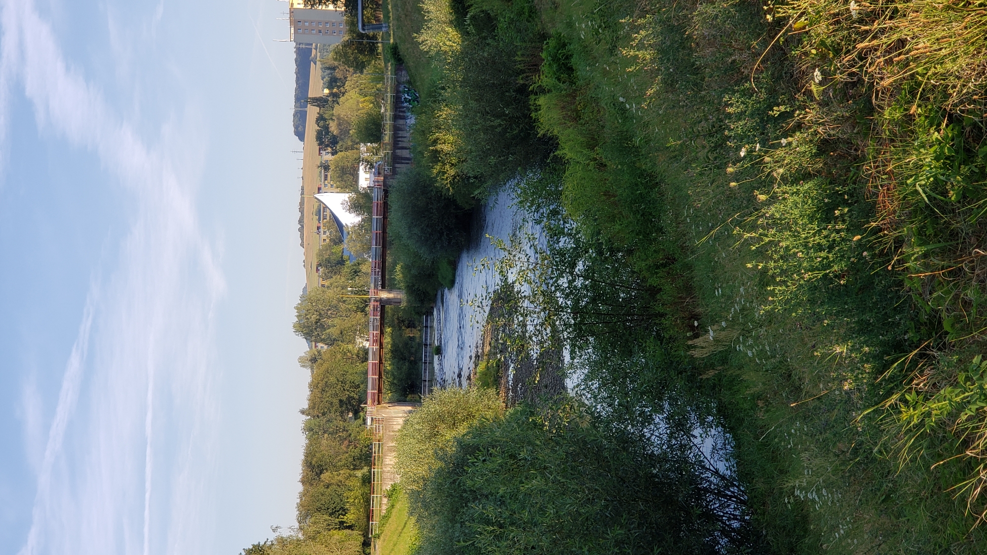 The image shows a river flowing through a moderately urbanized area, likely Prešov, Slovakia. The river has visible rocky shallows and is flanked by lush green vegetation, including trees and bushes. A concrete bridge with a red railing spans the river in the middle ground, and beyond it, there are more trees and some buildings, including a white, modern-looking structure with a pointed roof and a tall yellow apartment building on the right. The sky is blue with some wispy white clouds. The overall impression is a natural landscape integrated into a developed environment, bathed in what appears to be late afternoon sunlight.