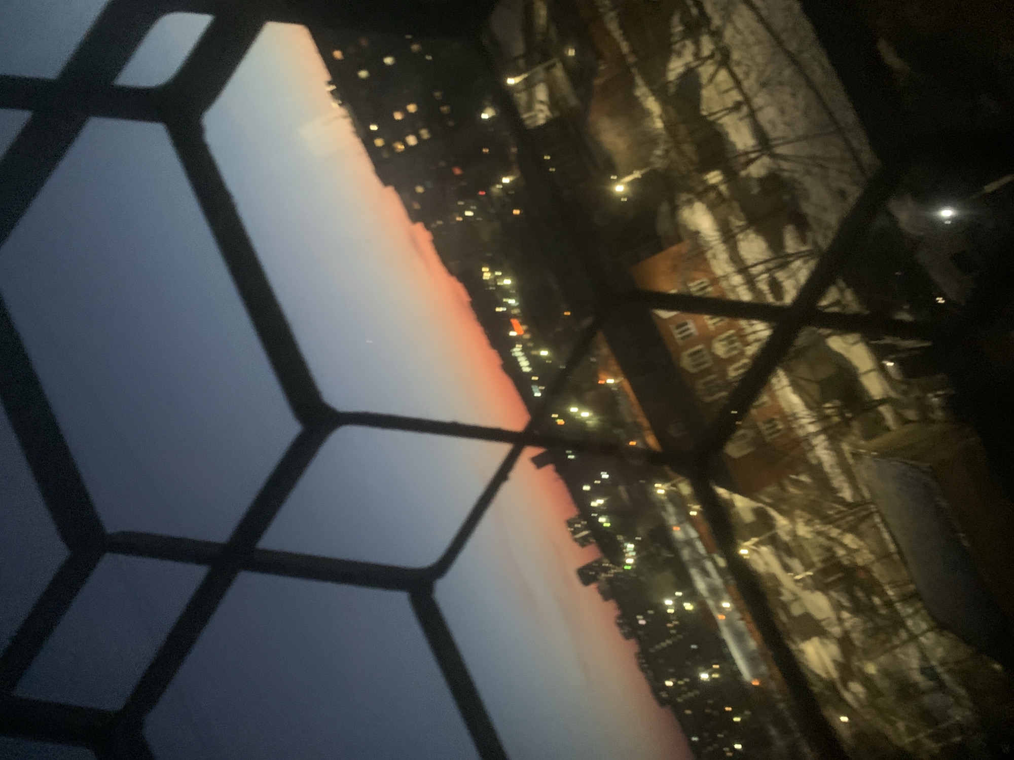The image shows a view of Izhevsk, Russia, at dusk or dawn, framed by a window with a multi-paned grid. The sky transitions from a dark blue to a pale orange or pink on the horizon. City lights are visible as small, bright dots across the horizon. Reflections in the window show a building covered in snow. There are no people visible.