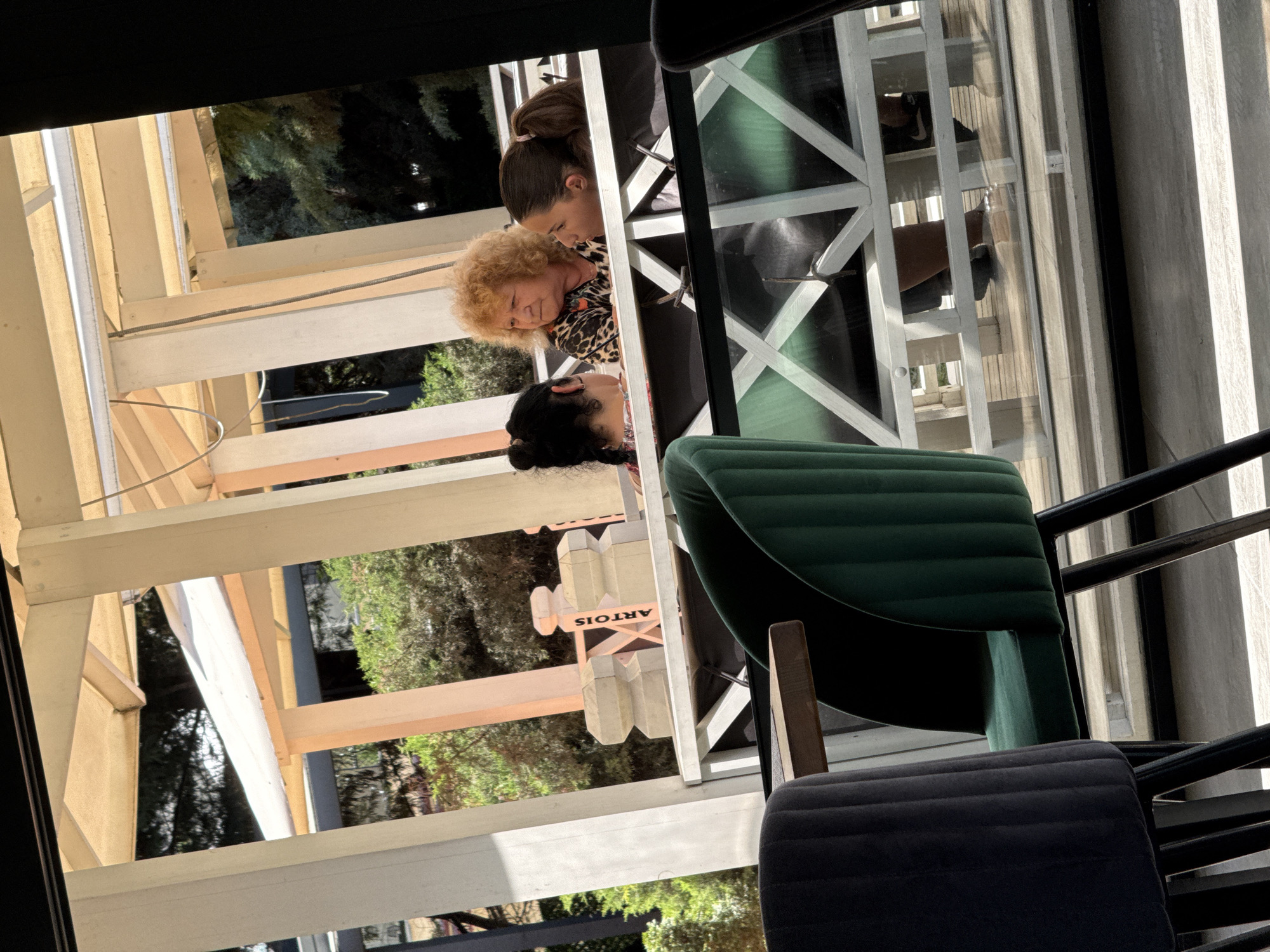 The image depicts an outdoor seating area, possibly a cafe or restaurant, in Shumen, Bulgaria. Three individuals are seated at a table, viewed from behind and through a reflective surface. In the foreground, two plush chairs are visible, one dark grey and the other emerald green, with dark metal legs. Beyond these chairs, the subjects are seated at a white railing. The individual on the left has dark hair styled in a bun and is wearing a patterned top. The person in the center has light-colored, curly hair and is wearing a leopard-print top. The individual on the right has dark hair tied in a ponytail and is looking towards the person in the center. A white railing with a crosshatch pattern separates the seating area from a lower level. Behind the railing, there are architectural elements including cream and pale orange support columns and a tan awning. The background shows greenery, suggesting an outdoor setting. A sign with the word "ARTOIS" is visible on the white railing. The overall lighting suggests daytime.