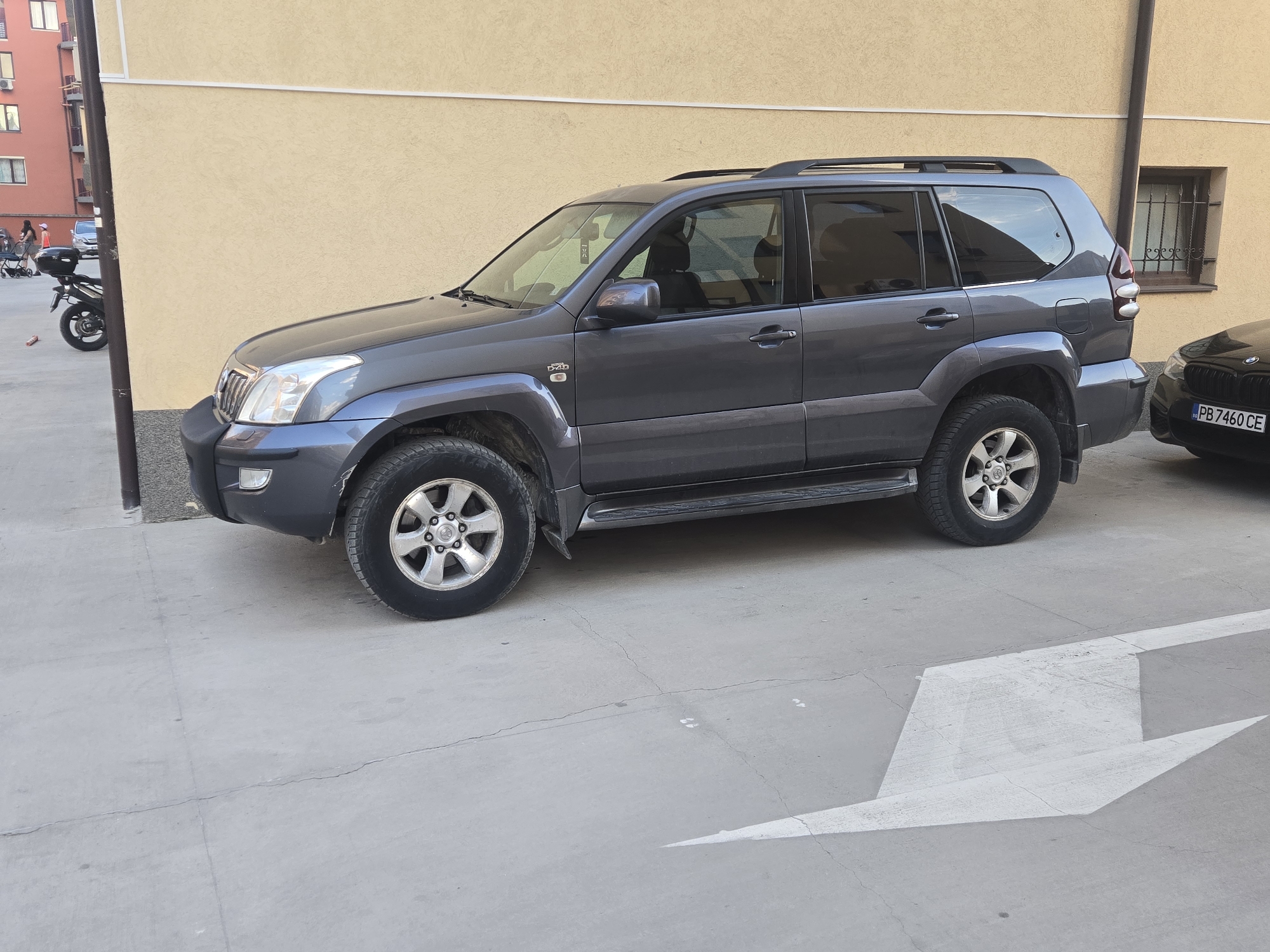 A dark gray Toyota Land Cruiser Prado is parked on a concrete surface with a large white painted arrow pointing to the right, next to a light yellow building in an outdoor urban setting, possibly in the city of Unknown, Unknown. A black motorcycle and other vehicles are visible in the background.