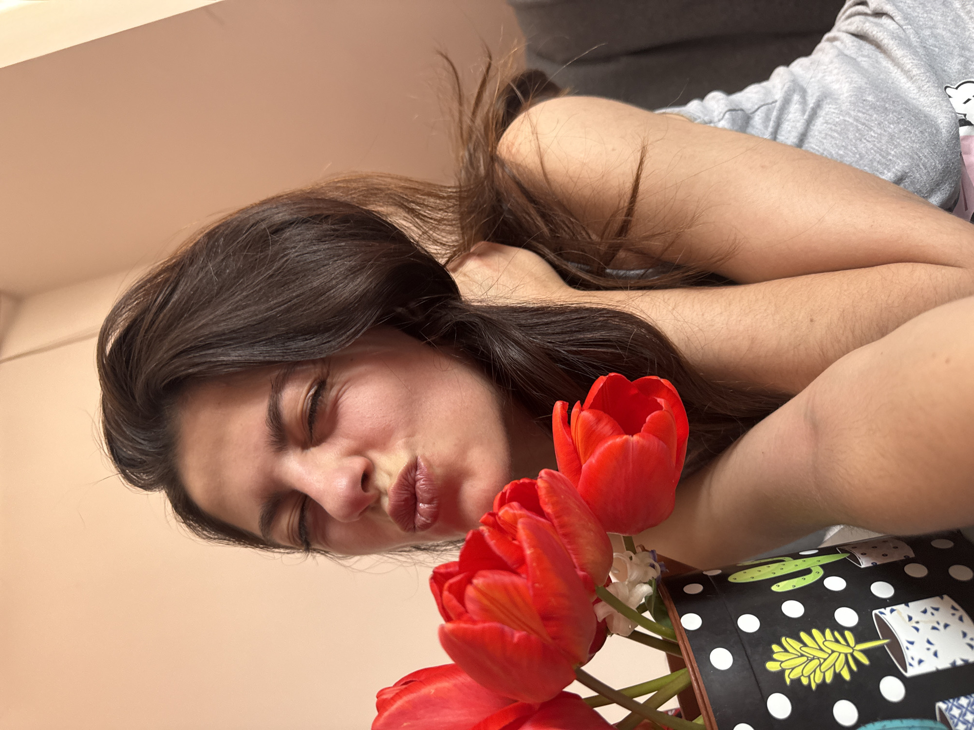 A young woman is lying down with a bouquet of red tulips. She is wearing a gray shirt and has brown hair. The bouquet of flowers is in a black box with a cactus and plant pot design. The environment is indoors, with a light-colored wall in the background. The location is not discernible from the image and cannot be determined as Plovdiv, Bulgaria.