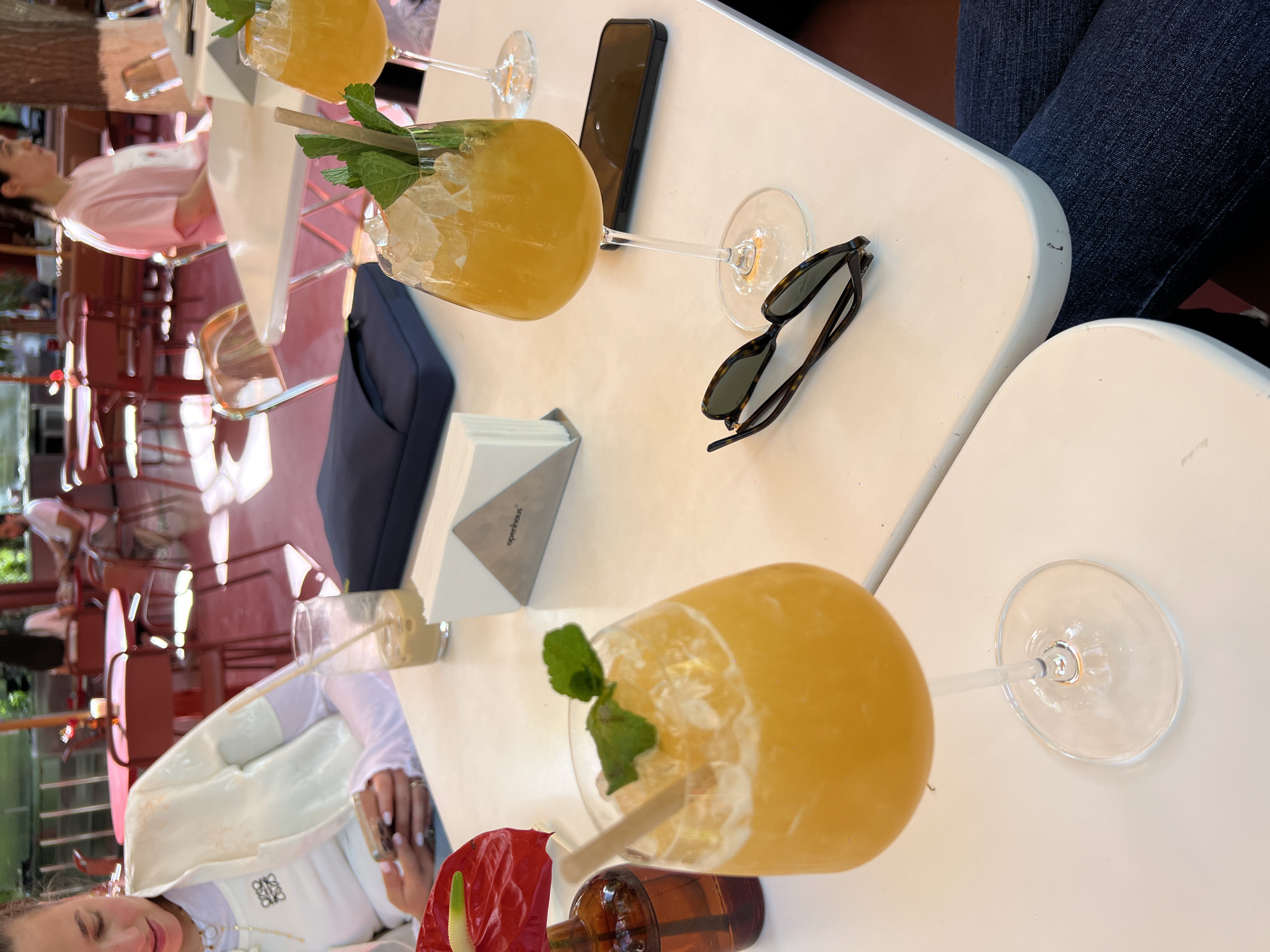 The image captures a scene at an outdoor seating area of a likely restaurant or bar in an unidentified city. Several cocktails, garnished with mint and possibly fruit, are placed on white tables. The angle is taken from above the table top, showing two people seated at separate tables. One person on the left is partially visible, wearing a white top with decorative gold detailing. The person's skin is fair, and they have manicured nails. There is a single red flower on the table next to them and another cocktail in a glass. On the right side of the photo, a person wears dark blue jeans. Between them, a phone and a pair of sunglasses rest on the table. The surrounding environment includes reddish-brown patio furniture and a glimpse of trees and structures beyond, suggesting the setting is in a garden or outdoor area in a residential area of the city.