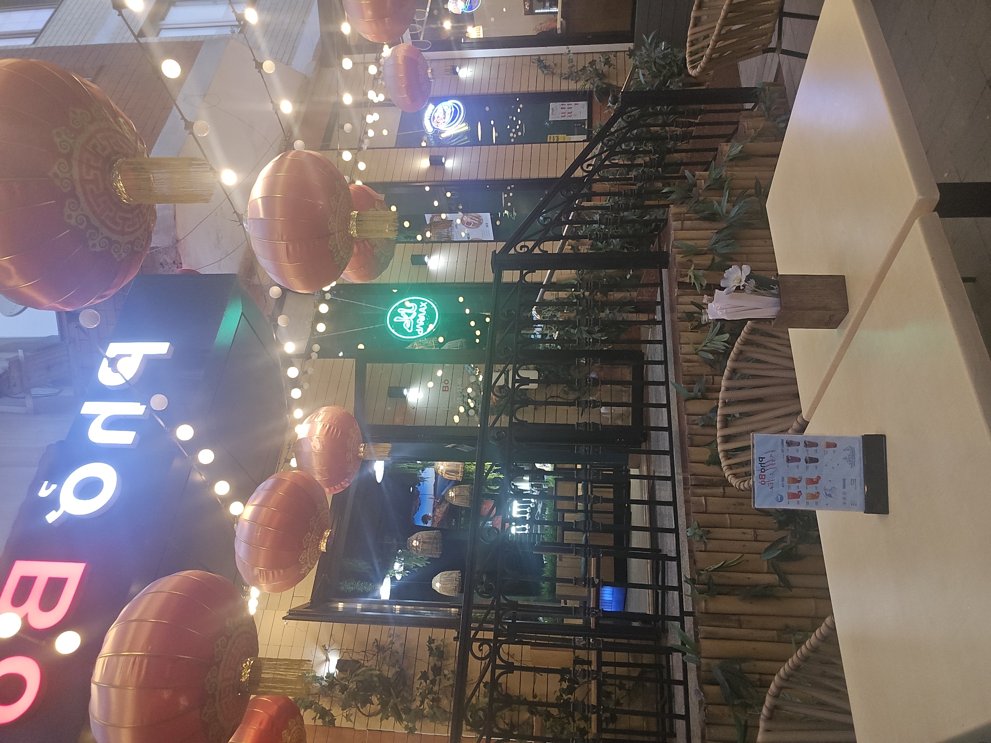 Here is a description of the image:
The image shows an outdoor seating area of a restaurant or cafe, likely at night, judging by the lighting. The main focal points are the large red lanterns hanging above, typical of Asian-themed establishments. The restaurant's name is "bo bo" in large neon letters. The outdoor area is enclosed by a black metal railing, with bamboo-like decor behind it. Tables and chairs are visible, and a menu stand sits on one of the tables. The overall impression is that this is a vibrant, inviting spot. The city and location are unknown.