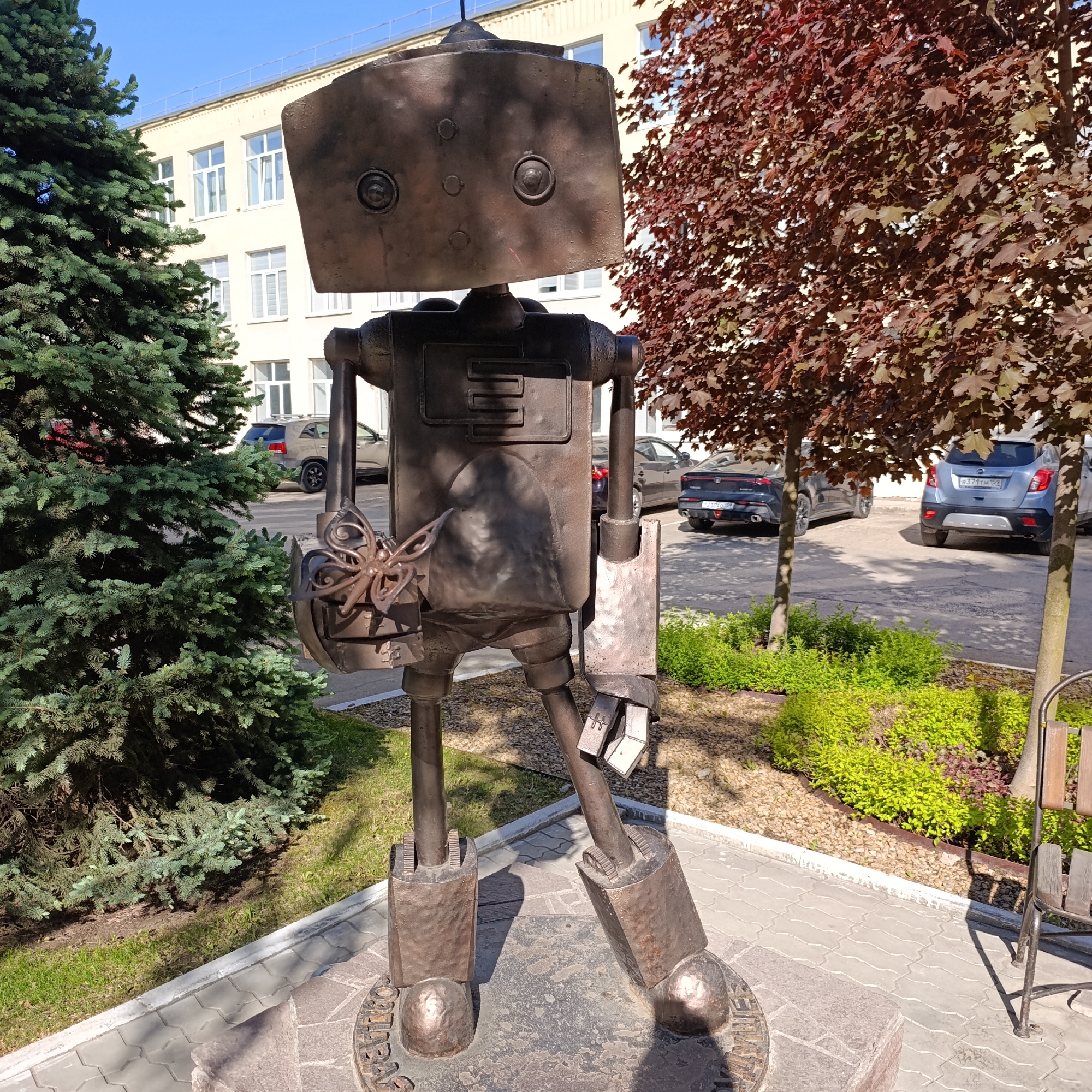 The image shows a metal robot statue standing on a circular base, located outdoors in Chaikovsky, Russia. The robot has a rectangular head with two round "eyes" and other geometric features. It has a boxy body with arms and legs, and holds a butterfly-shaped object in its left hand. It stands on a gray stone platform surrounded by grass, a green pine tree on the left, and a red-leafed tree on the right. In the background is a light-colored building with windows and parked cars.